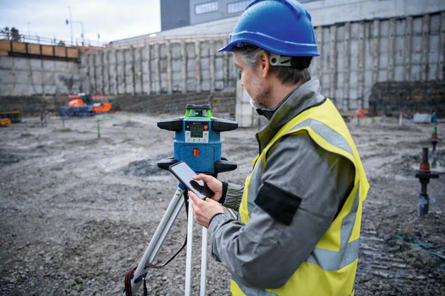 Ein Bauarbeiter mit blauem Helm und gelber Weste benutzt ein Gerät auf einem Stativ. Baustelle im Hintergrund.