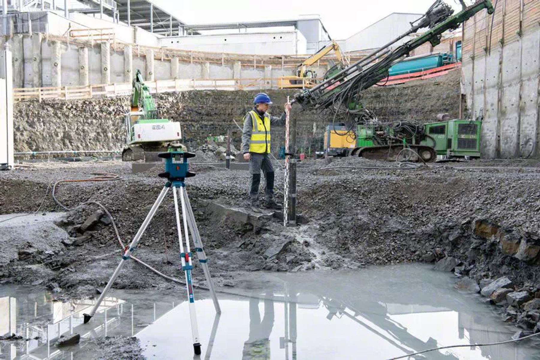 Baustelle mit Arbeiter, der eine Wasserwaage benutzt. Bagger und Bohrgeräte befinden sich im Hintergrund. Wasser und Kies befinden sich im Vordergrund.