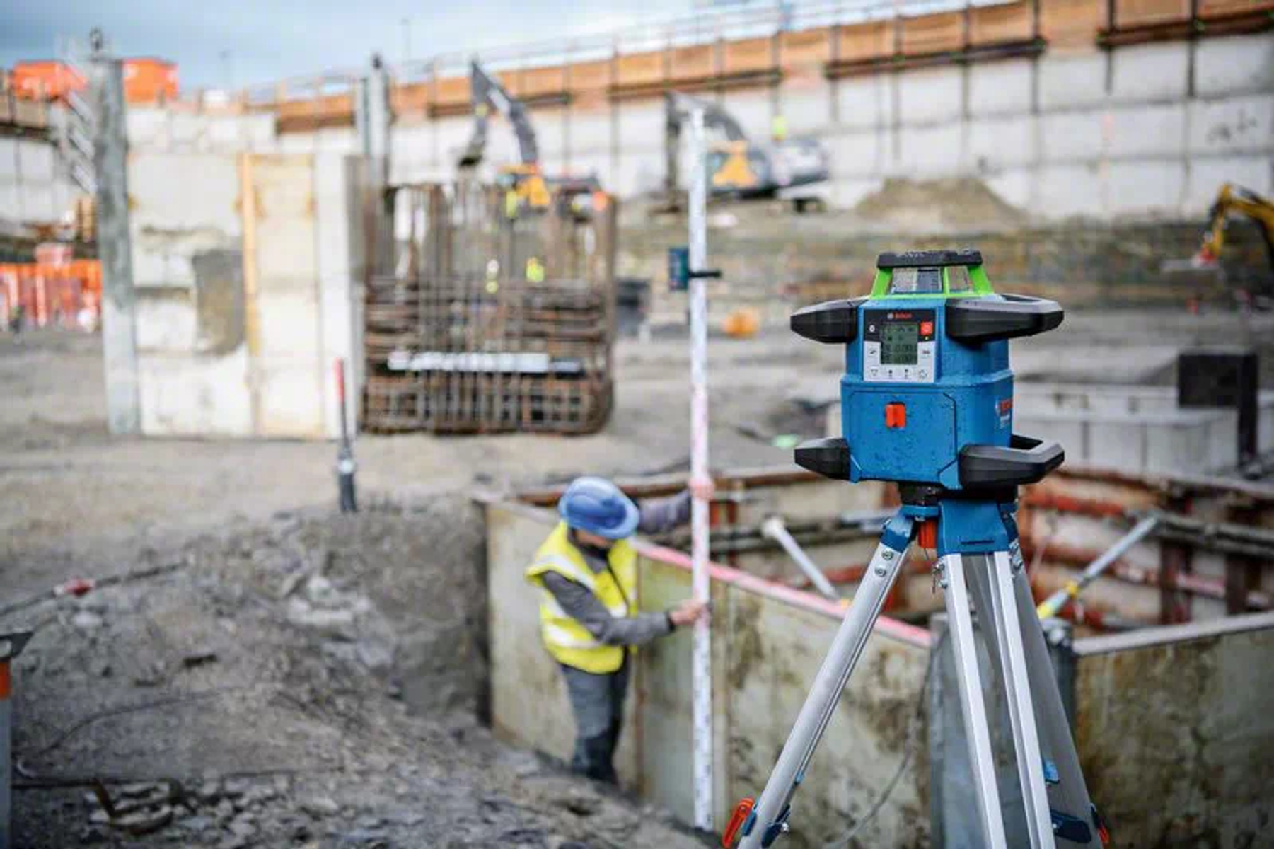 Baustelle mit blauem Laser-Nivelliergerät auf einem Stativ. Ein Arbeiter in einer Weste benutzt einen Messstab. Im Hintergrund befinden sich Maschinen.