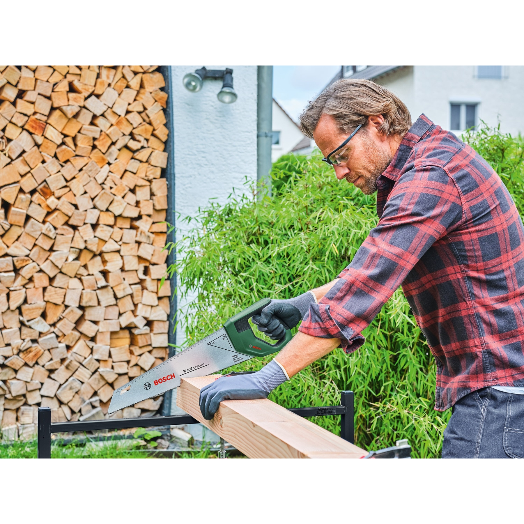 Ein Mann sägt im Freien ein Holzbrett. Er trägt eine Schutzbrille und ein rot kariertes Hemd. Im Hintergrund ein Holzhaufen.