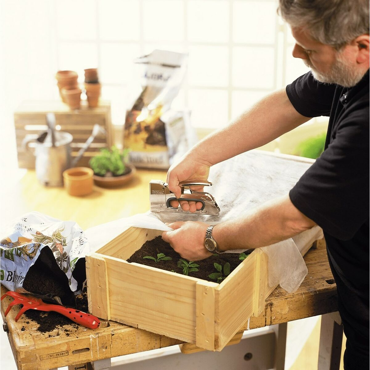 Ein Mann heftet Stoff über eine Holzkiste mit Setzlingen. Werkzeuge und Erdsäcke liegen auf einer Werkbank. Hintergrund Töpfe und Pflanzen.