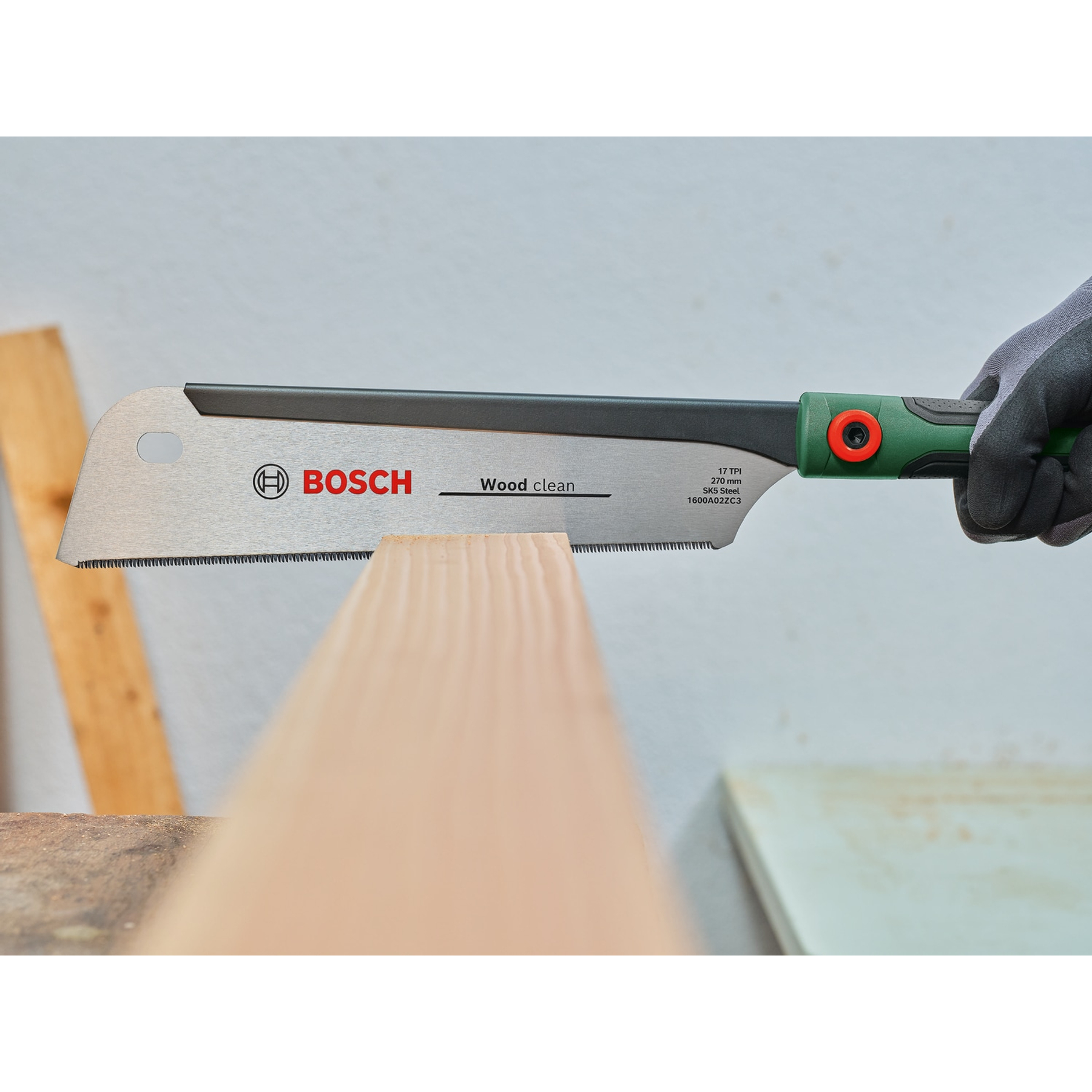 Eine Hand hält eine Bosch-Holzsäge und schneidet ein Holzbrett. Die Säge ist grün und schwarz. Der Hintergrund ist eine Werkstatt.