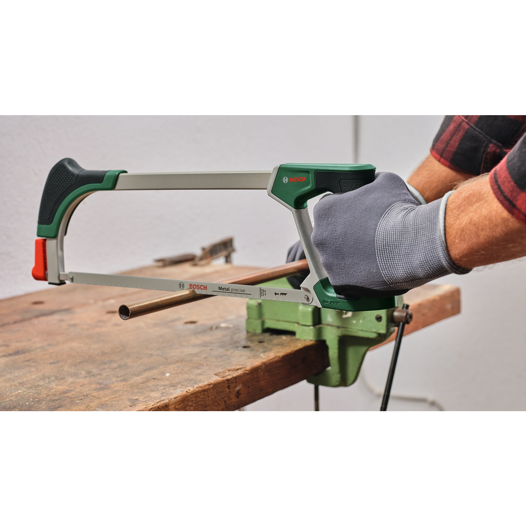 Eine Person schneidet mit einer Bosch-Säge ein Kupferrohr. Die Säge ist grün und grau. Hände tragen graue Handschuhe. Werkbank.