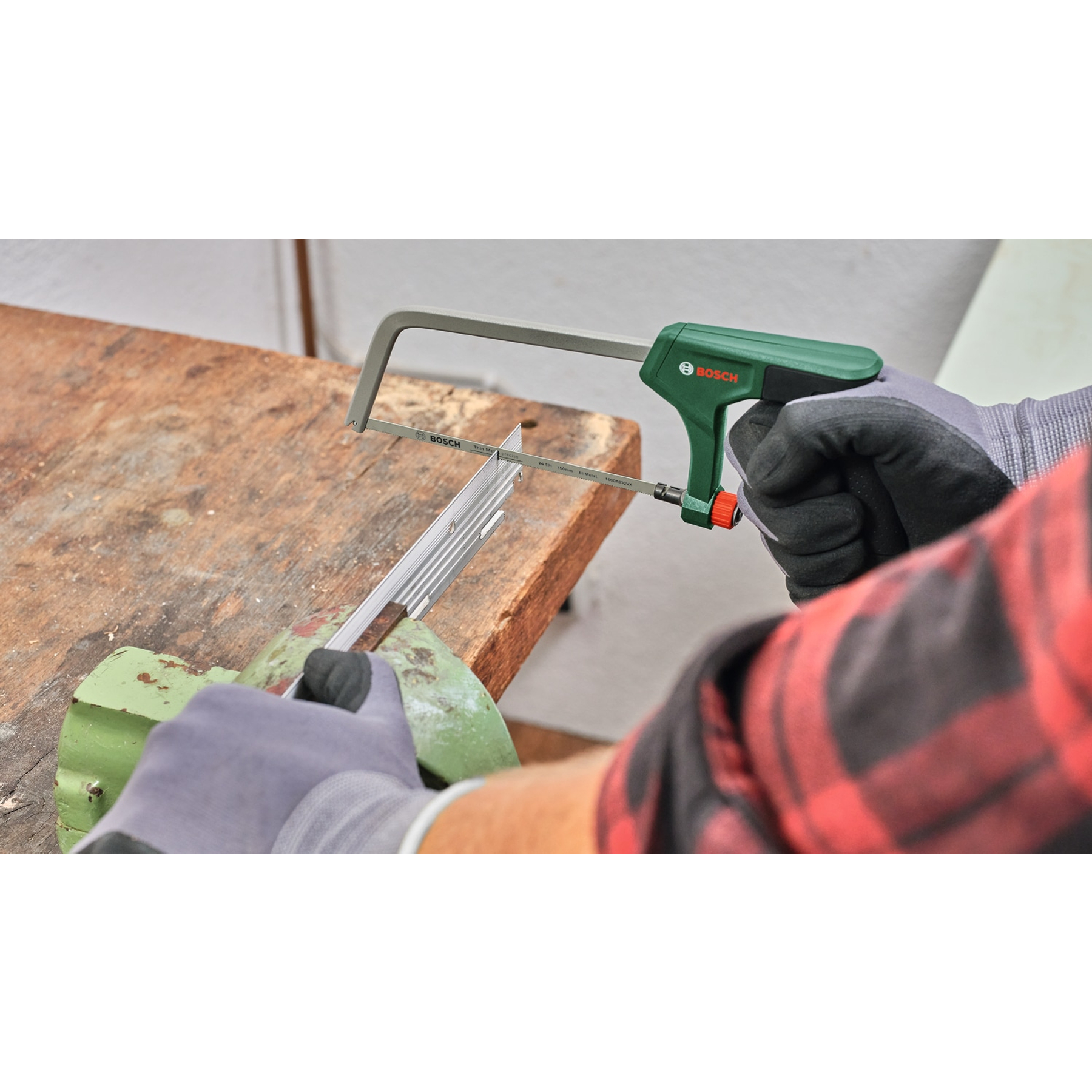 Una persona sta segando una barra di metallo con un seghetto Bosch verde e grigio. Sono visibili il tavolo di legno e il morsetto di metallo verde.