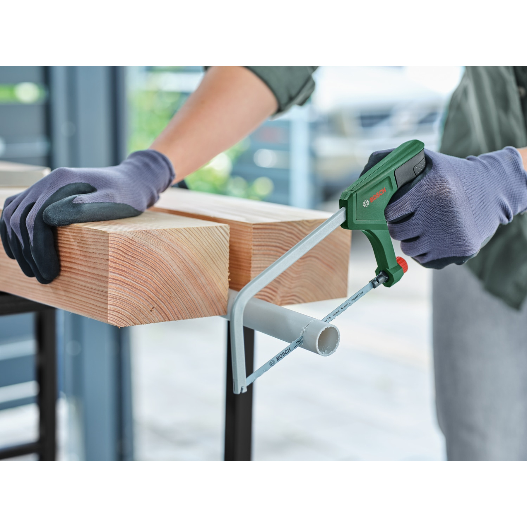 Una persona usa un seghetto alternativo Bosch verde per tagliare un tubo bianco, con guanti, su una superficie di legno.