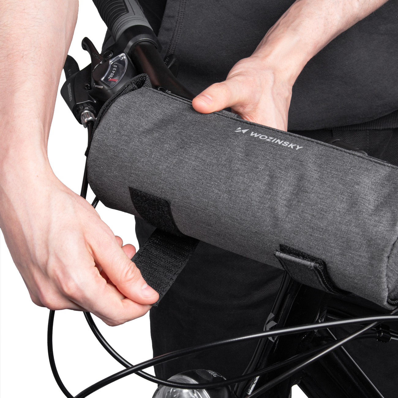 Eine Person befestigt eine graue Fahrradtasche an einem schwarzen Fahrradlenker. Die Tasche hat einen Klettverschluss und den Markennamen 'Wozinsky'.