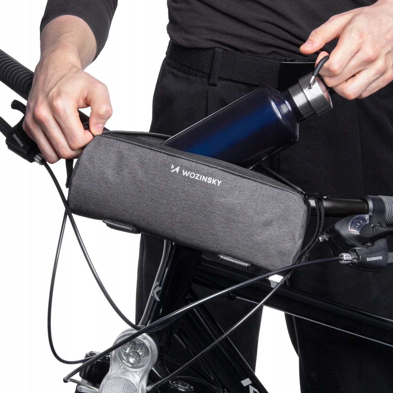 Eine Person legt eine Wasserflasche in eine graue Fahrradtasche. Das Fahrrad ist schwarz. Die Tasche hat die Marke Wozinsky.