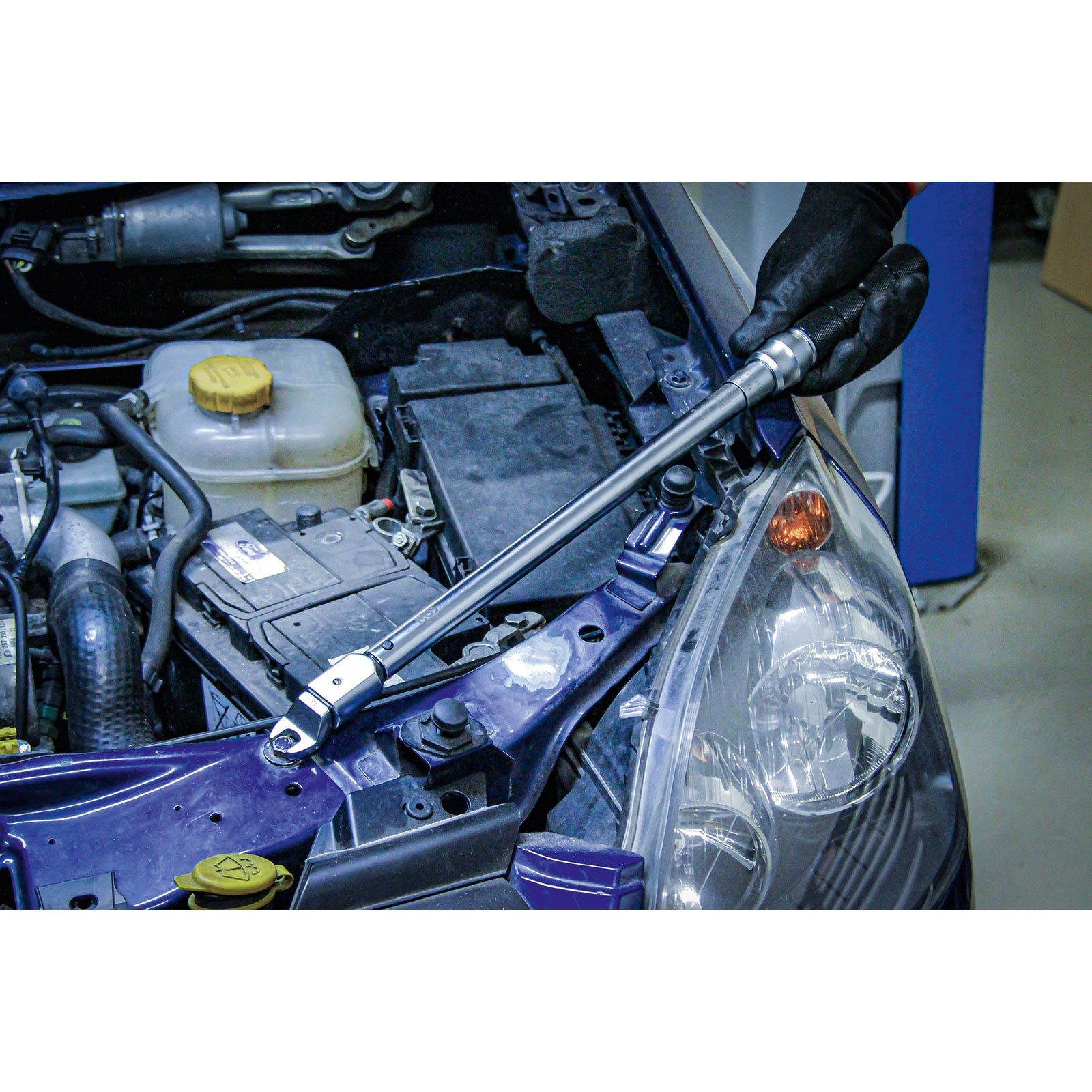 Ein Mechaniker mit schwarzen Handschuhen benutzt einen Schraubenschlüssel an einem Automotor. Die offene Motorhaube zeigt Motorteile und einen Scheinwerfer.
