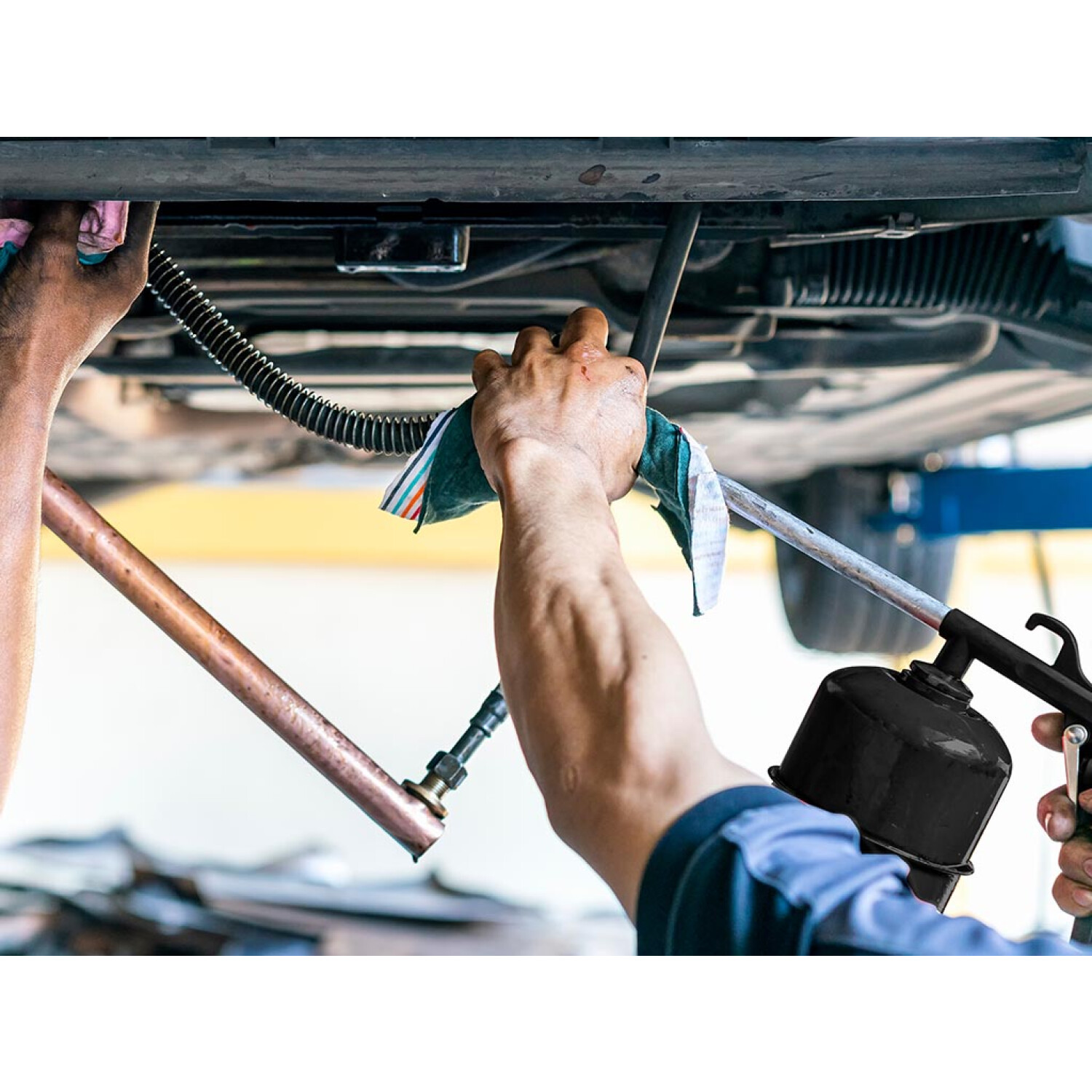Ein Mechaniker arbeitet unter einem Auto und benutzt ein Werkzeug. Hände sind zu sehen, einige halten einen Lappen, andere eine schwarze Spritzpistole.