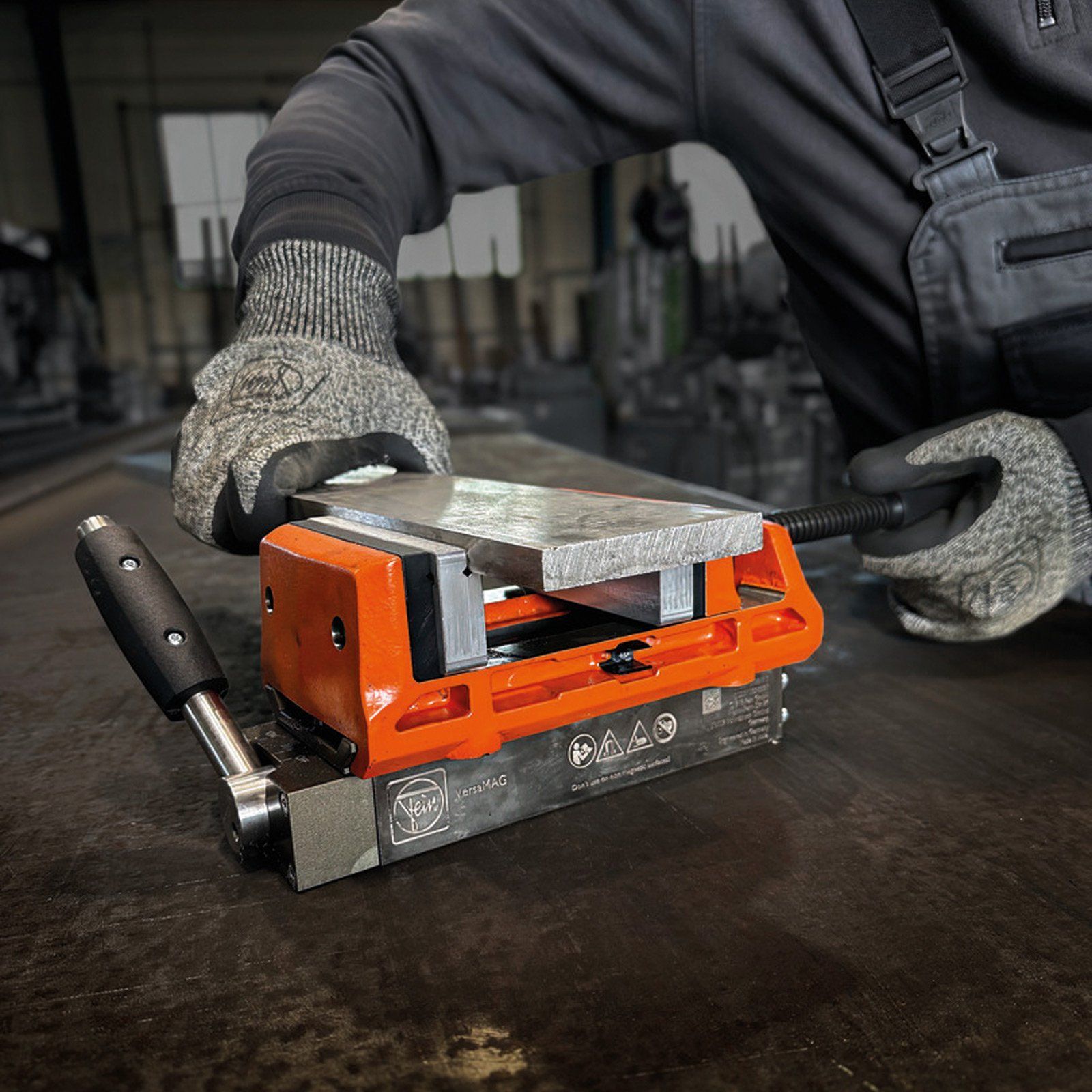Eine Person in Handschuhen bedient einen orangefarbenen Metallschraubstock und hält eine Metallplatte auf dunkler Oberfläche.