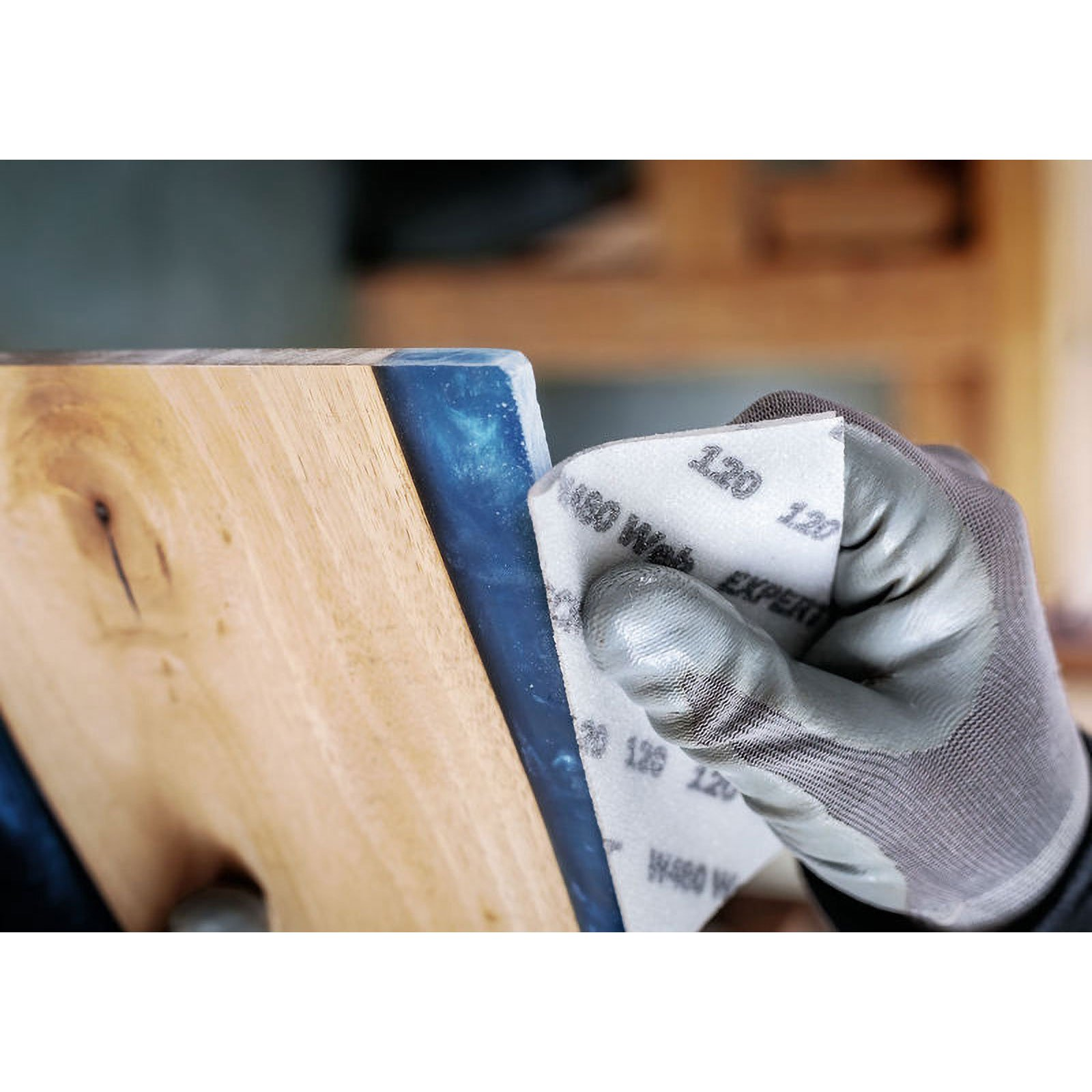 Una mano guantata sta carteggiando una superficie di legno con bordi in resina blu, usando un blocco abrasivo 120. Primo piano.