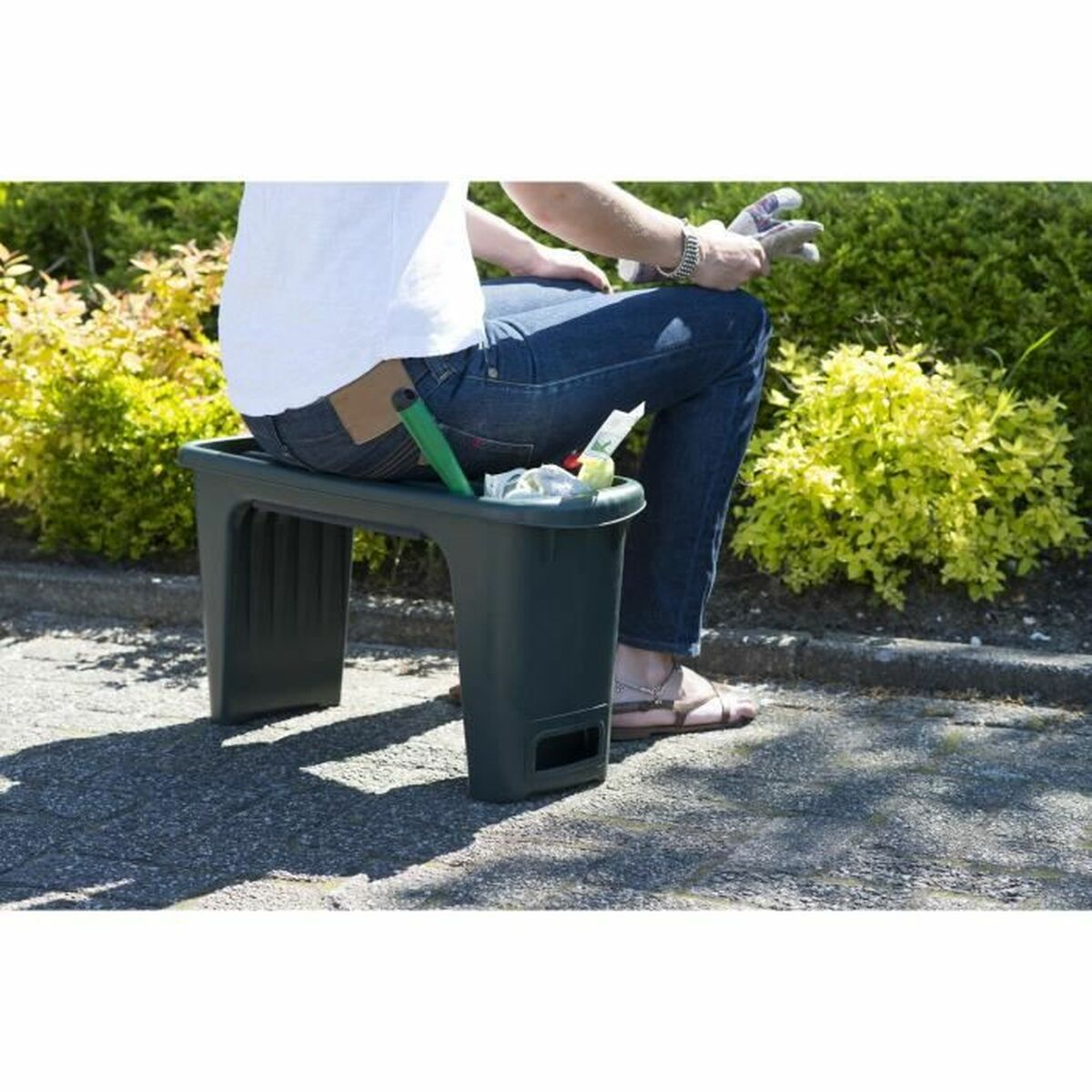 Eine Person sitzt auf einem dunkelgrünen Gartenhocker und hält Handschuhe. Im Tablett des Hockers befinden sich Werkzeuge. Büsche und eine gepflasterte Fläche sind im Hintergrund.