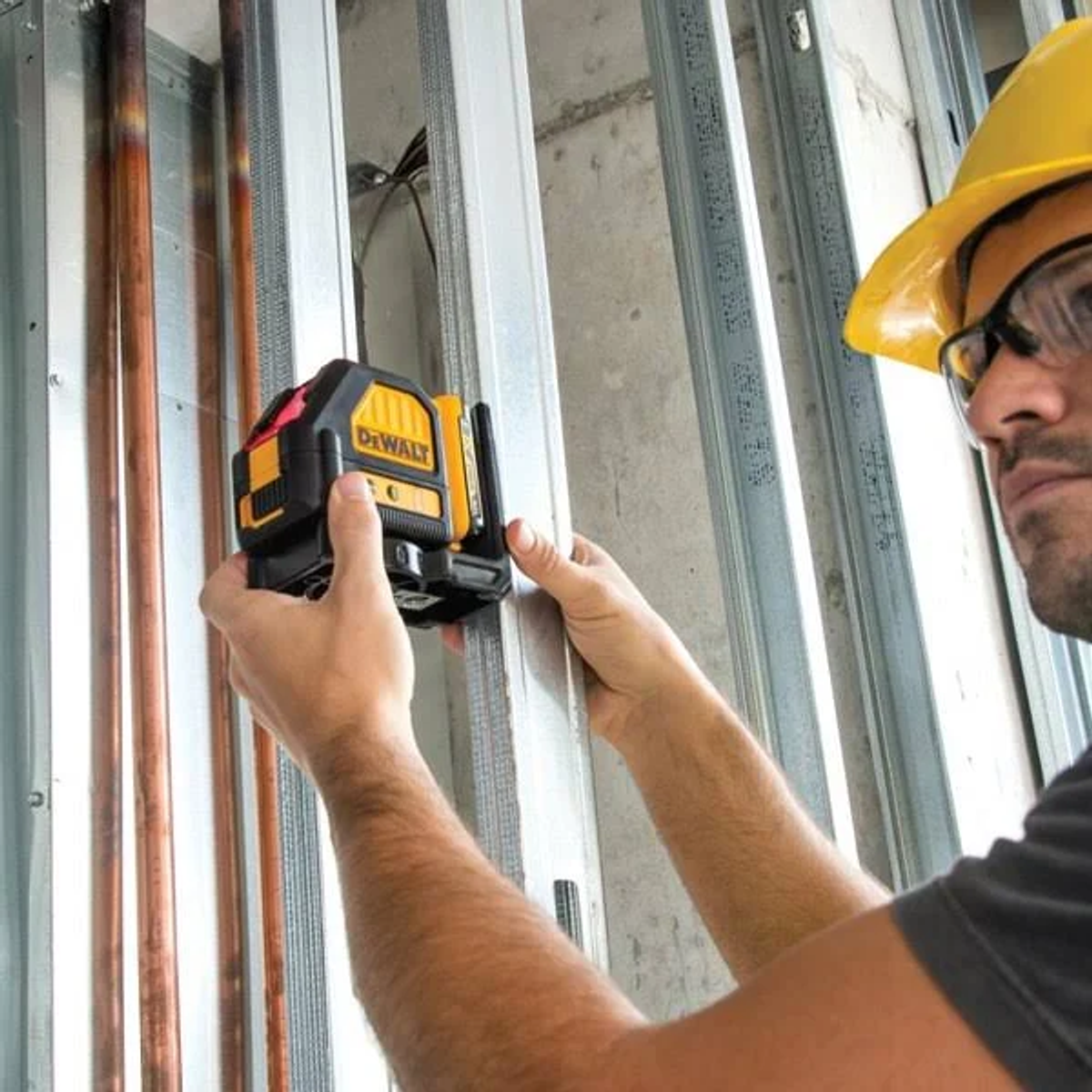 Ein Bauarbeiter mit gelbem Helm benutzt einen Laser-Wasserwaage. Kupferrohre und Metallstifte sind im Hintergrund.