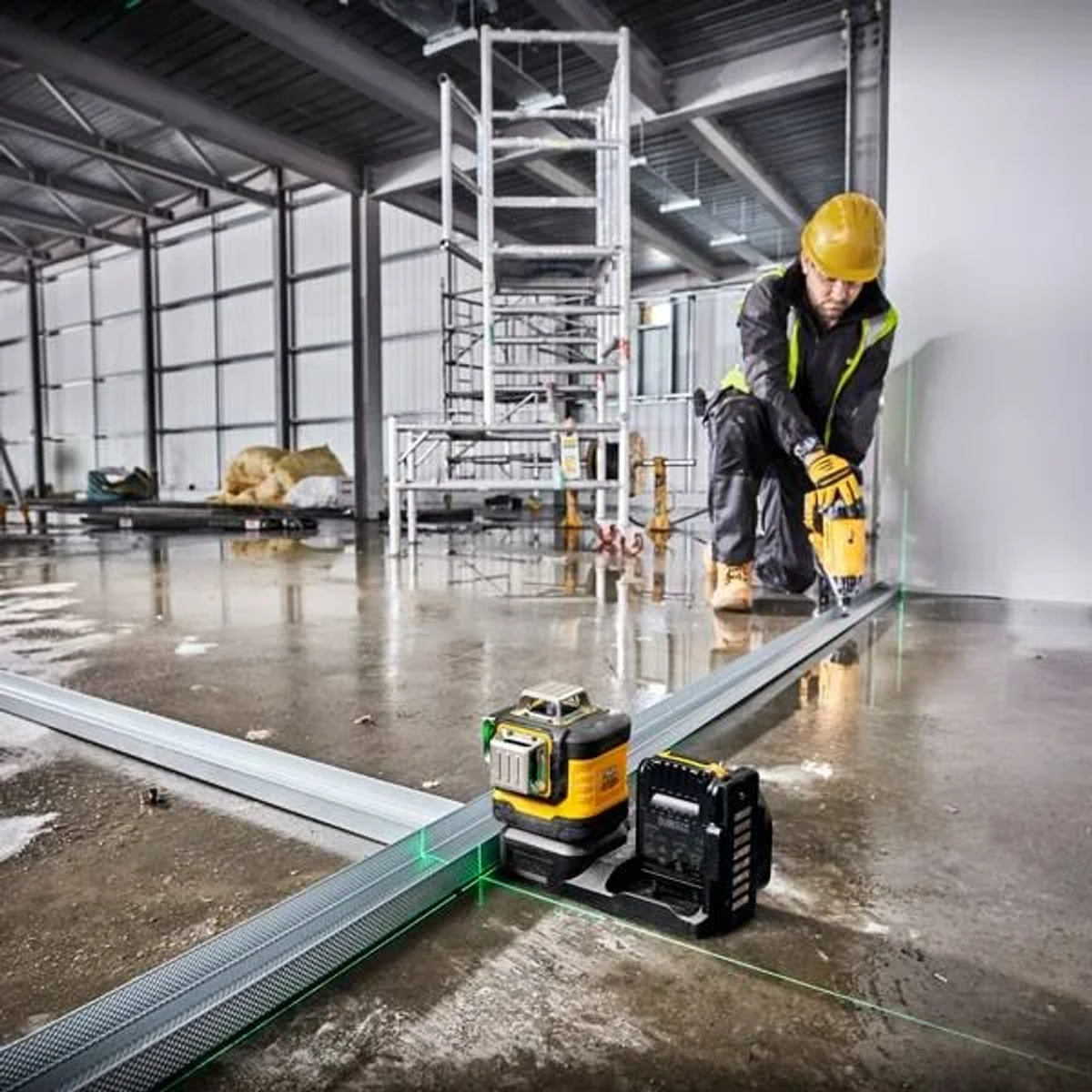 Ein Arbeiter mit gelbem Helm verwendet einen Laser am Metallträger. Baustelle mit Gerüst und Werkzeugen im Hintergrund.