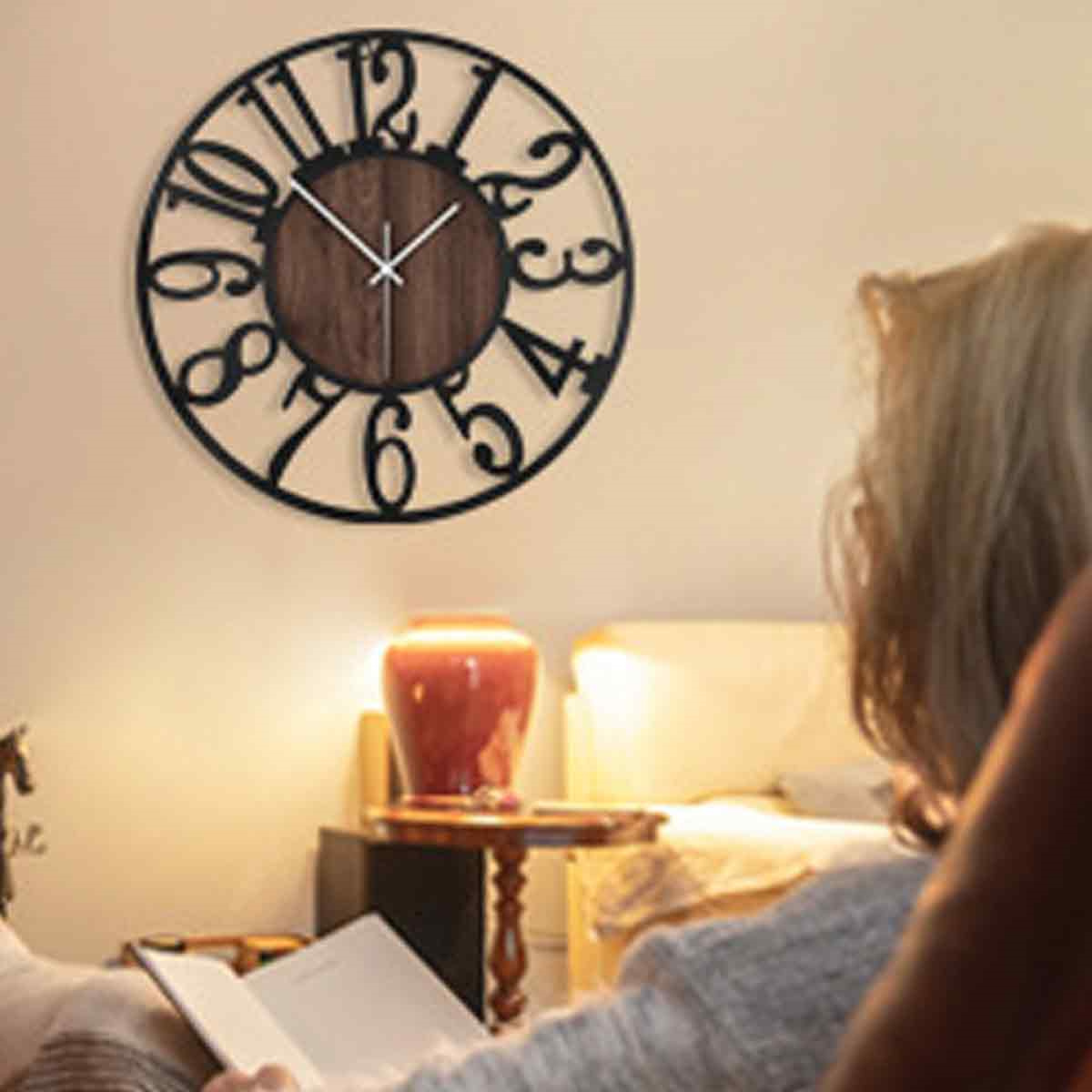 Eine schwarze Uhr hängt an einer Wand. Eine Frau sitzt mit einem Buch. Eine Lampe steht auf einem Tisch.
