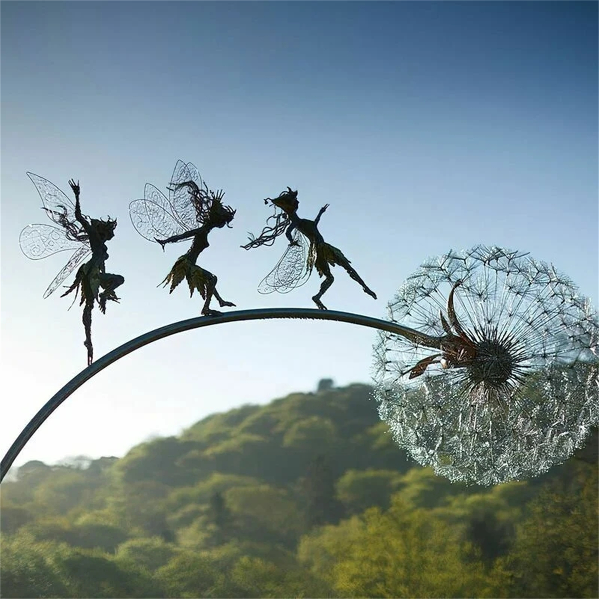 Drei Feen-Skulpturen auf einem gebogenen Metallstab in der Nähe einer Pusteblume. Grüne Hügel und blauer Himmel im Hintergrund.