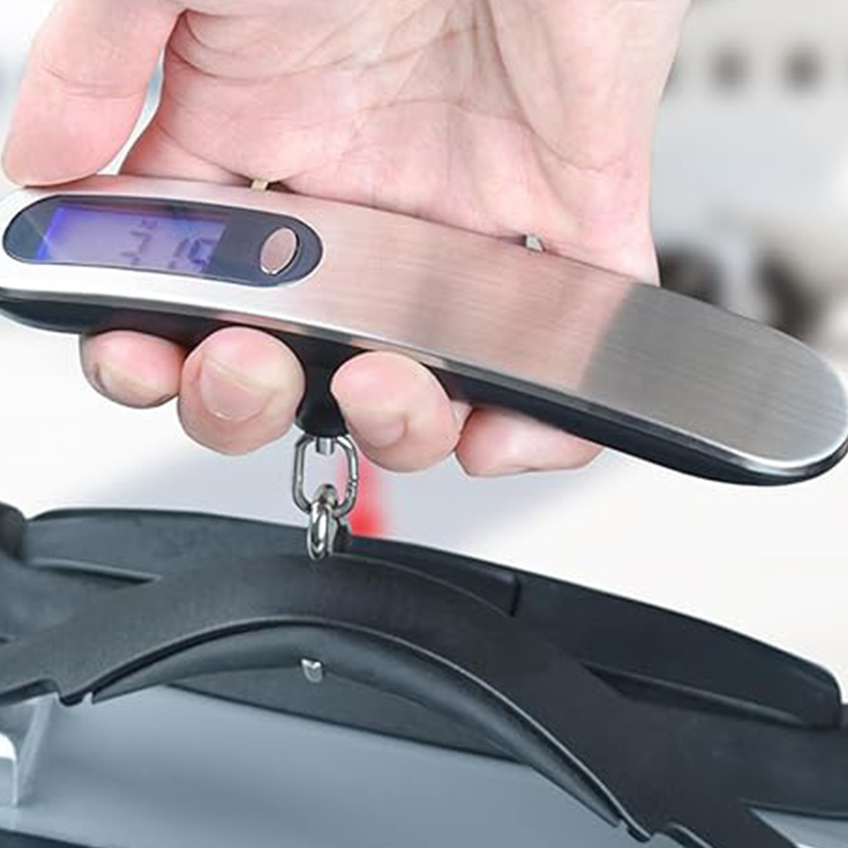 Hand hält eine silberne Gepäckwaage mit digitaler Anzeige. Darunter ist ein schwarzes Objekt befestigt.