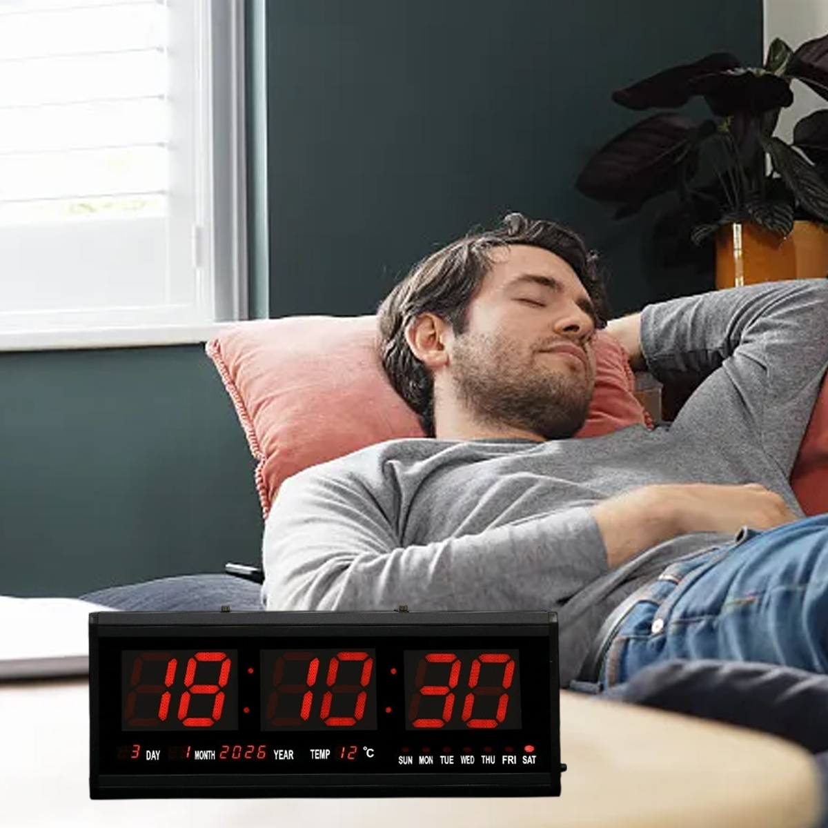 Ein Mann schläft auf einer Couch. Eine Digitaluhr zeigt 18:10:30. Datum und Temperatur werden ebenfalls angezeigt. Der Hintergrund hat ein Fenster.