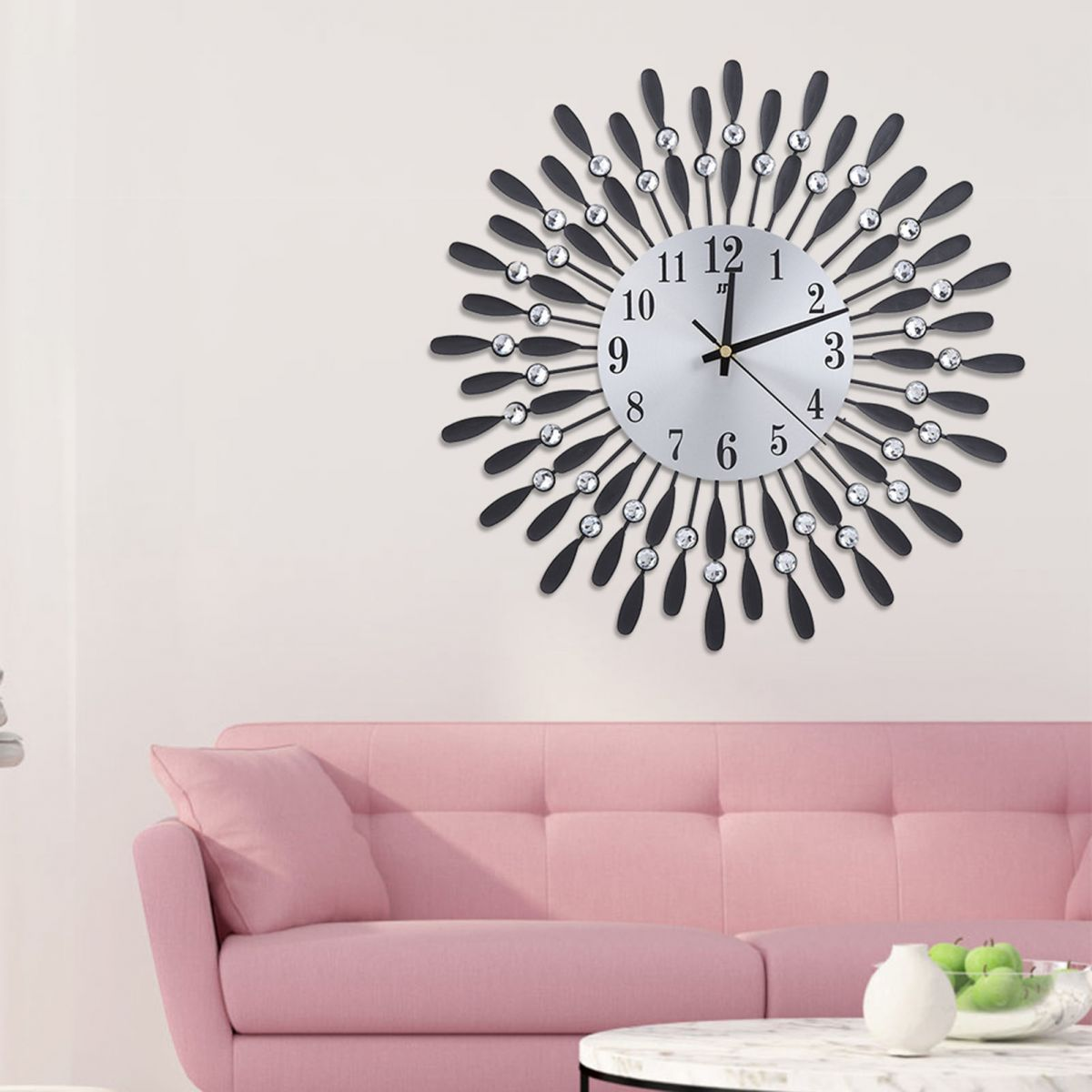 Eine dekorative Wanduhr mit schwarzen Akzenten und Kristallen an einer weißen Wand. Darunter befindet sich ein rosa Sofa. Ein Tisch mit Früchten.