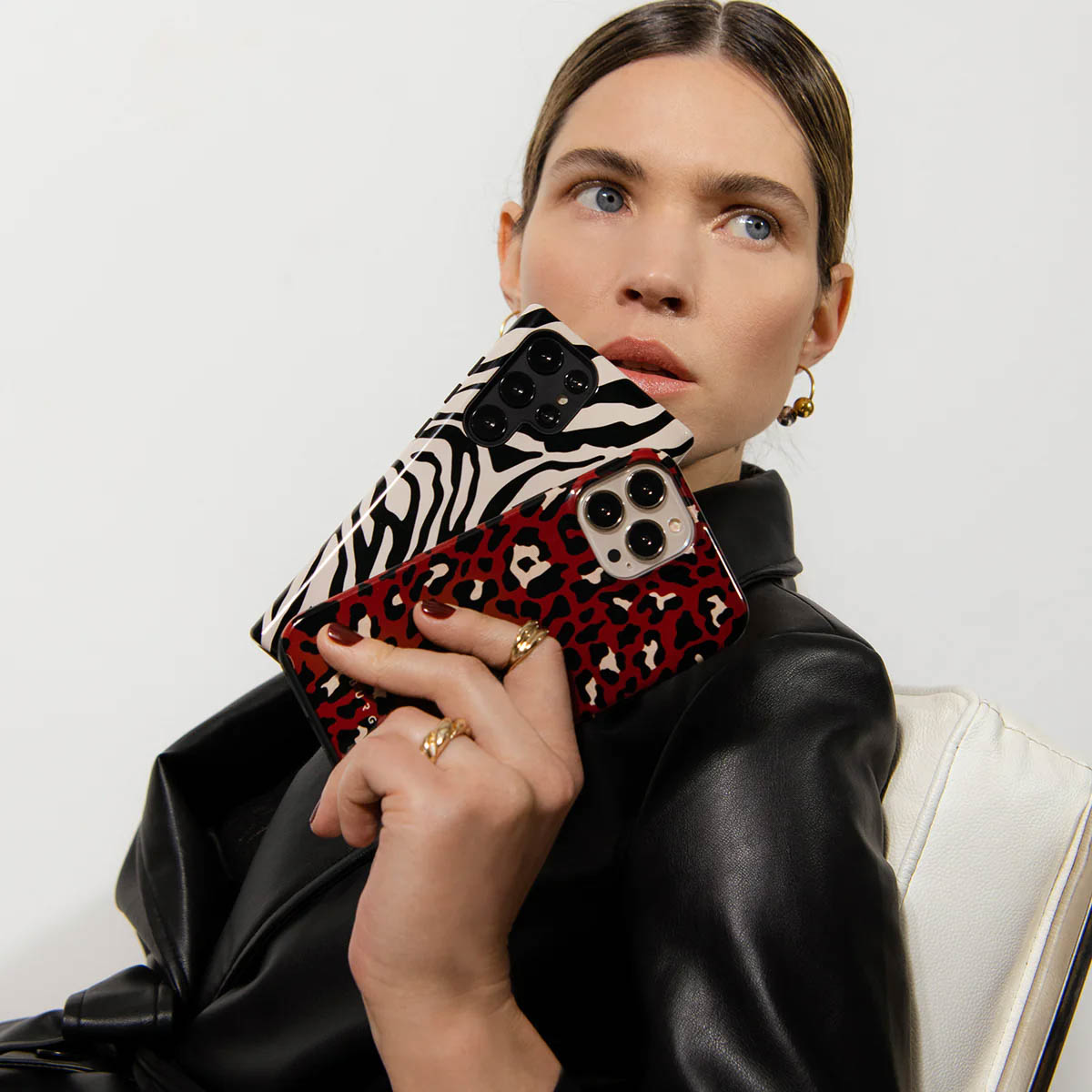 Een vrouw houdt twee telefoonhoesjes vast. Eén heeft een zebraprint, de andere een luipaardprint. Ze draagt een zwarte jas en ringen.
