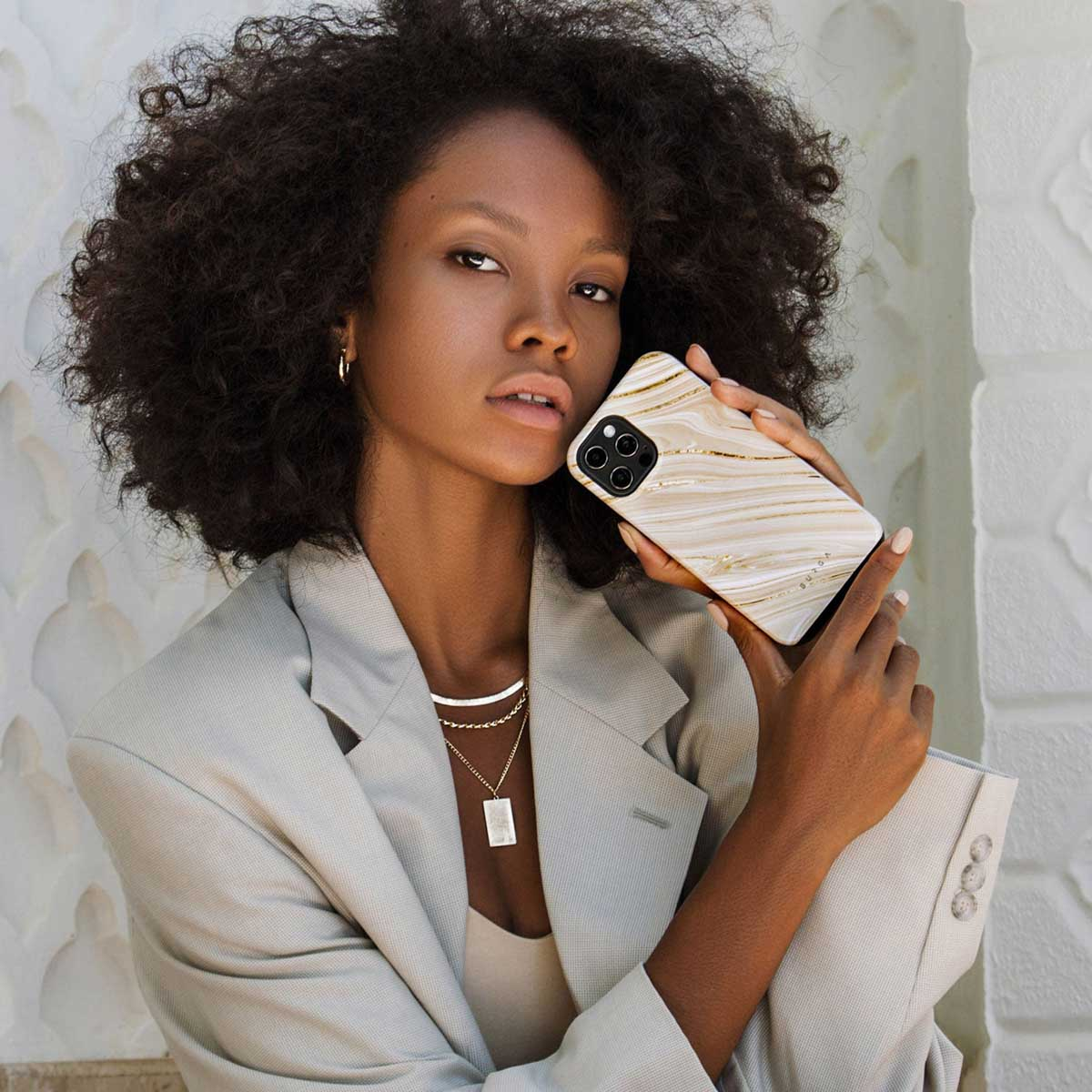 Une femme aux cheveux bouclés tient un téléphone avec un étui en marbre. Elle porte un blazer, un collier et des boucles d'oreilles, devant un mur blanc.