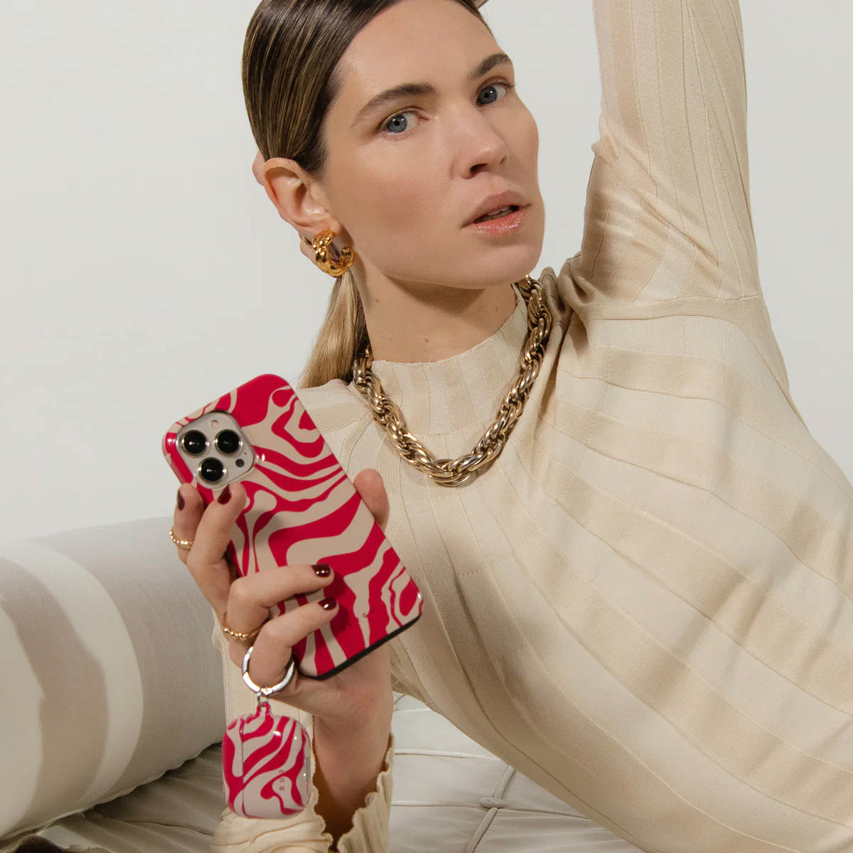 Vrouw houdt telefoon met rode zebra-hoes vast, draagt gouden sieraden en een beige trui, op een witte achtergrond.