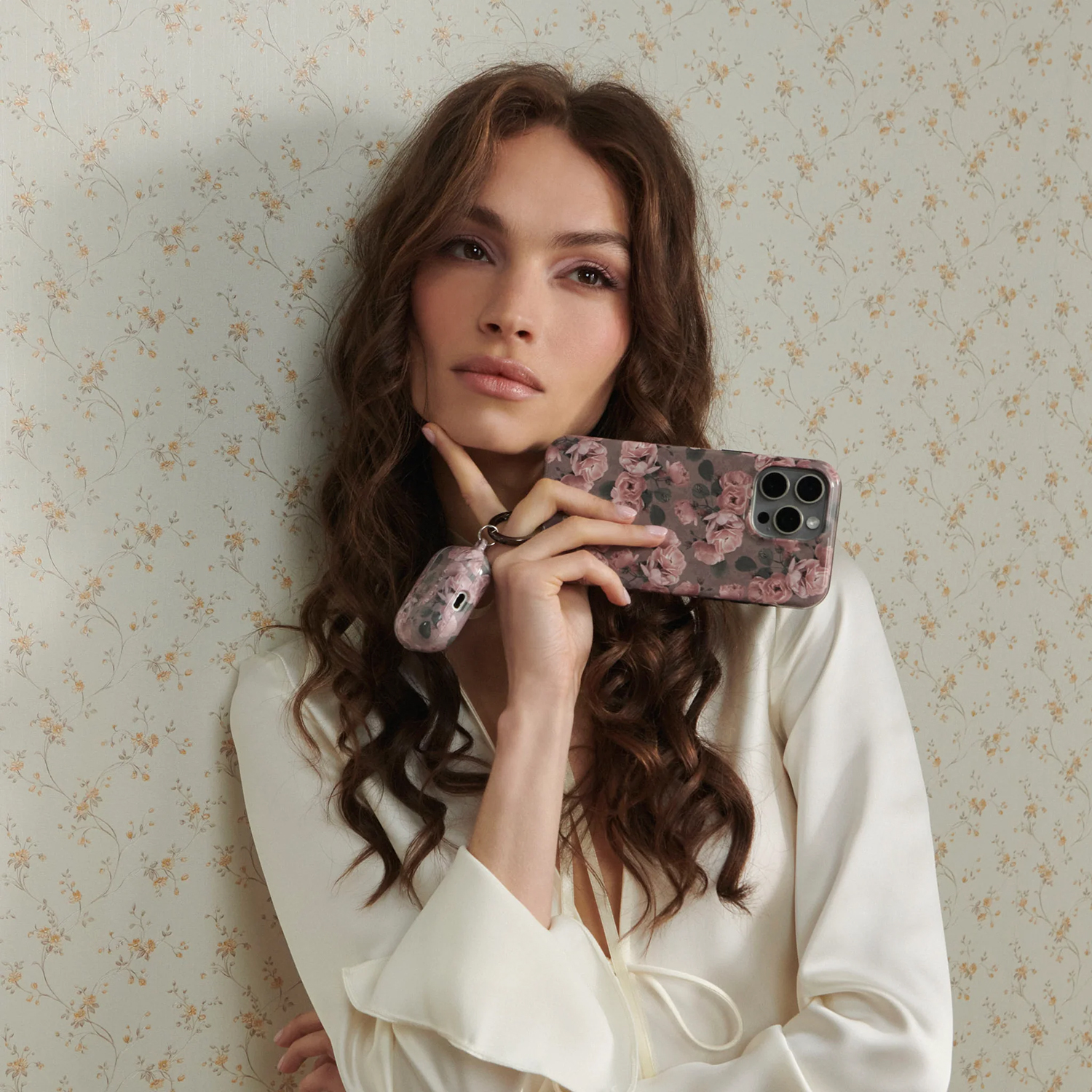 Vrouw houdt telefoon en oordopjescase vast, beide met bloemenpatroon. Ze draagt een wit overhemd, leunt tegen bloemenbehang en kijkt de camera in.