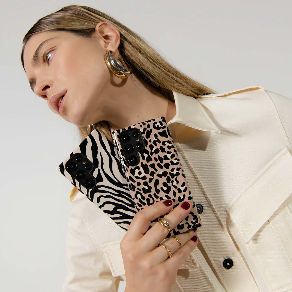 Een vrouw houdt twee telefoonhoesjes vast, een met zebraprint, de andere met luipaardprint, draagt gouden sieraden en een crèmekleurig jasje.