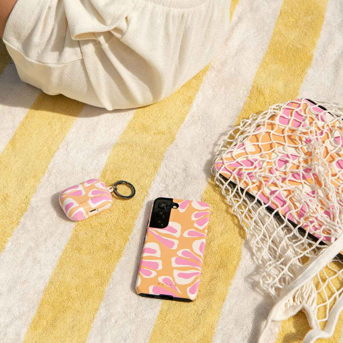 Een strandscène met een handdoek, telefoon, AirPods-hoesje en tas, allemaal met een roze en oranje patroon. Een persoon ontspant op de handdoek.