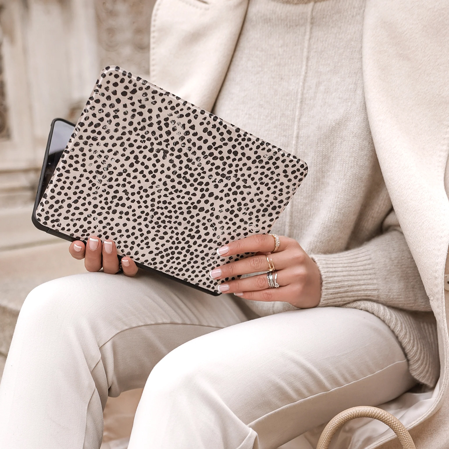 A person holds a tablet with a black and white spotted case. They wear a cream sweater, coat, and pants. Rings adorn their fingers.
