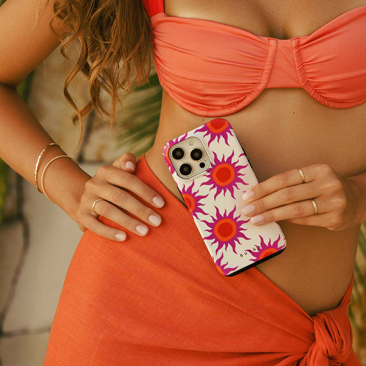 Een vrouw houdt een telefoon vast met een hoesje met zonnepatroon. Ze draagt een bikinitop en een oranje sarong. Gouden armbanden en ringen zijn zichtbaar.