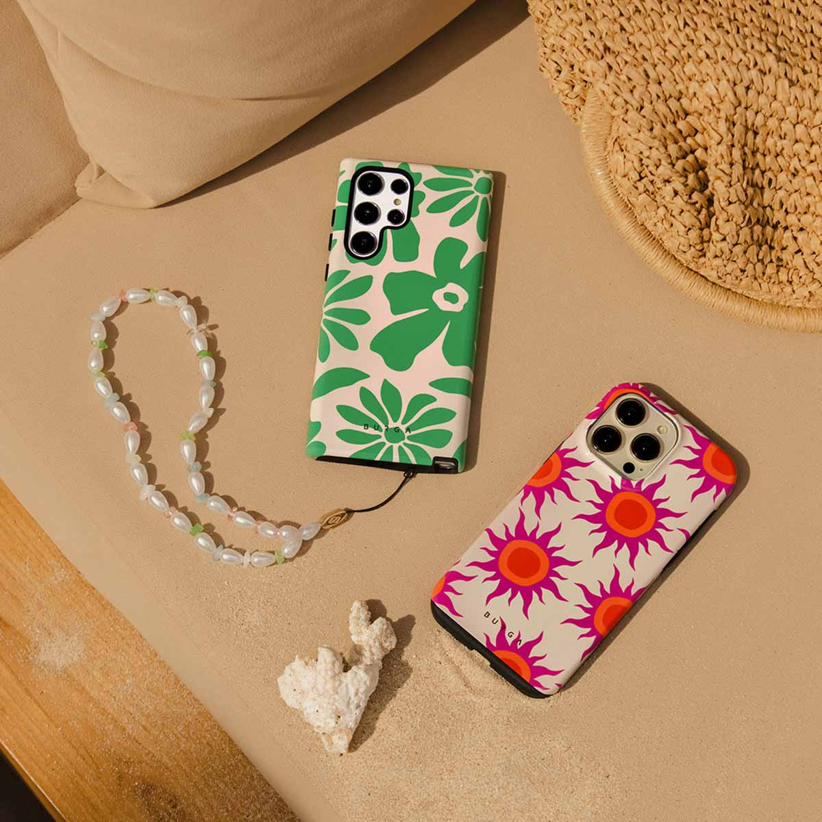 Twee telefoonhoesjes met bloemmotieven, een parelketting, een stuk koraal en een strohoed op een zanderige ondergrond.