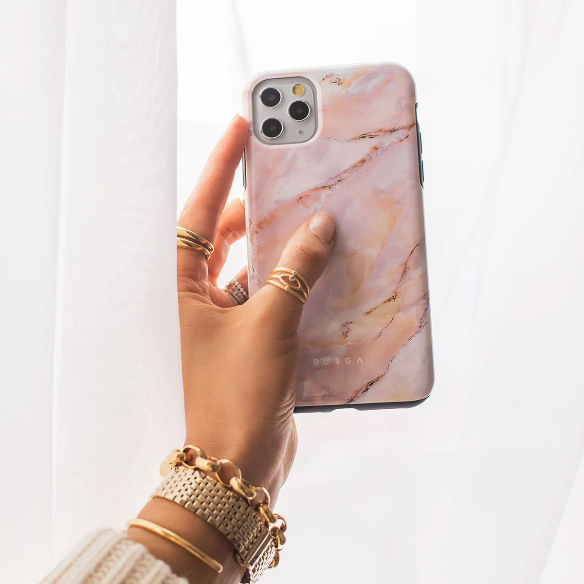 Een hand houdt een telefoon met een marmeren hoesje vast. Gouden ringen en een armband sieren de pols. Witte gordijn op de achtergrond.