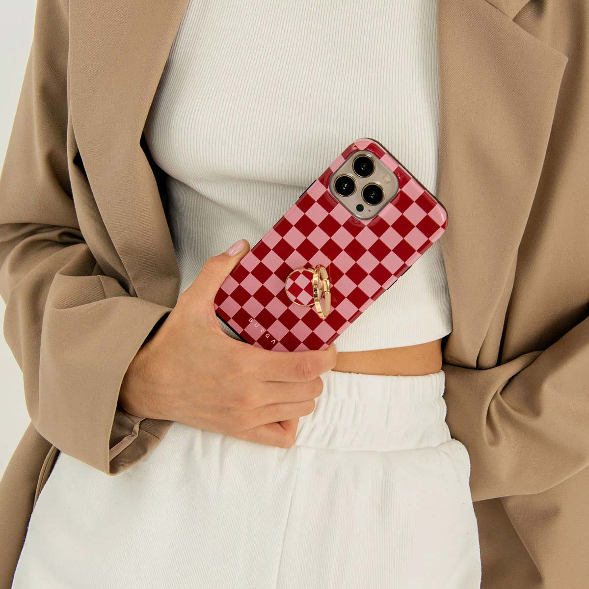 Een persoon houdt een telefoon vast met een rood-roze geruite hoes. Ze draagt een witte top, witte broek en een bruine blazer.