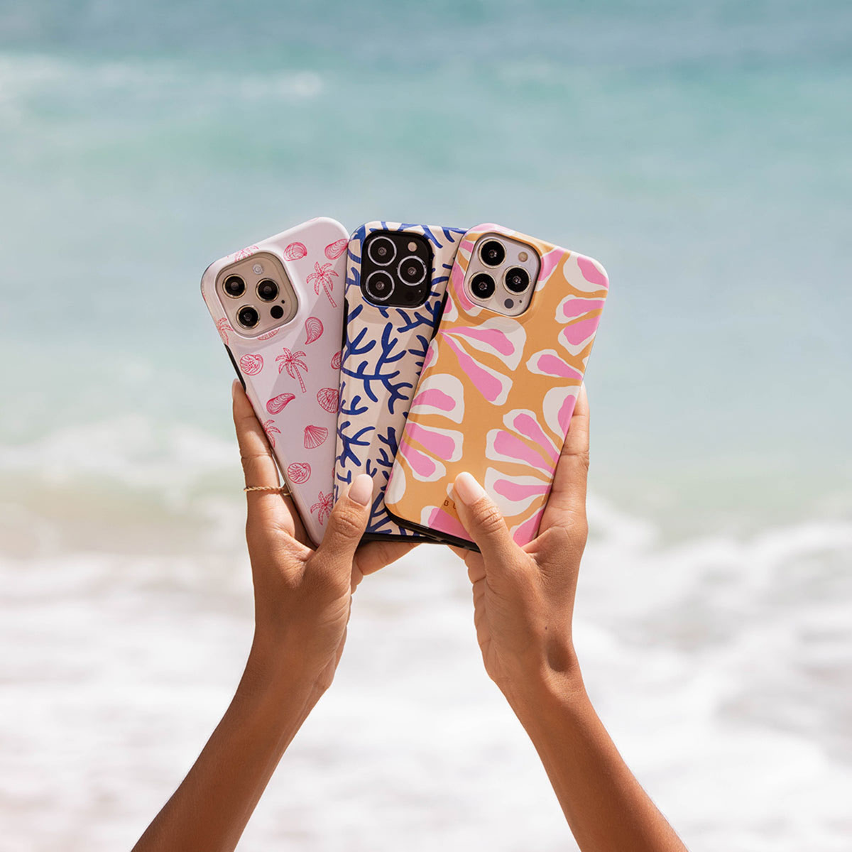 Hands holding three patterned phone cases against a blurred ocean backdrop. Cases feature coral, shell, and floral designs.