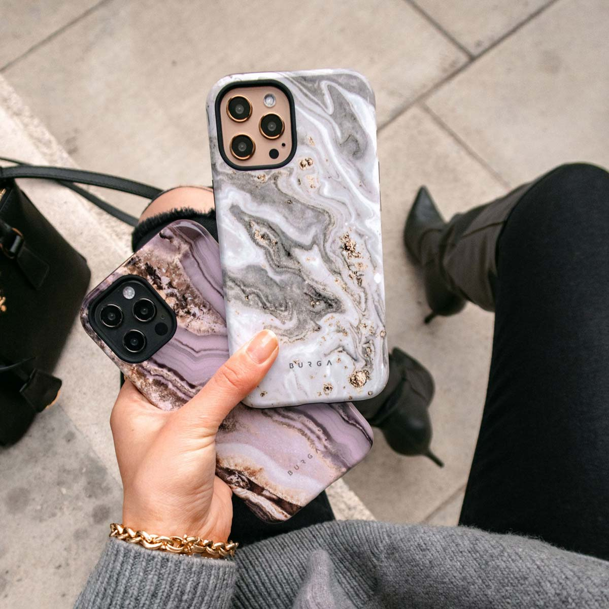 Een persoon houdt drie telefoonhoesjes met marmerontwerpen vast. Gouden armband om de pols. Zwarte laarzen en tas zijn zichtbaar op de achtergrond.