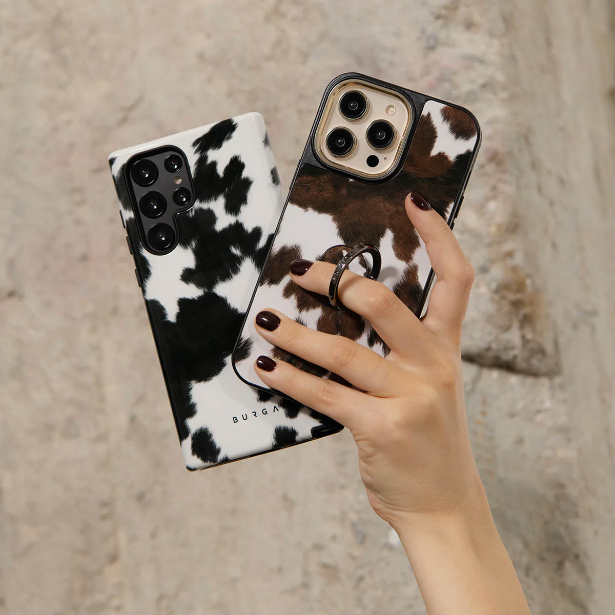 Een hand houdt een telefoon met een koeienprint hoesje vast. Een andere telefoon met koeienprint ligt ernaast. De achtergrond is getextureerd en licht.