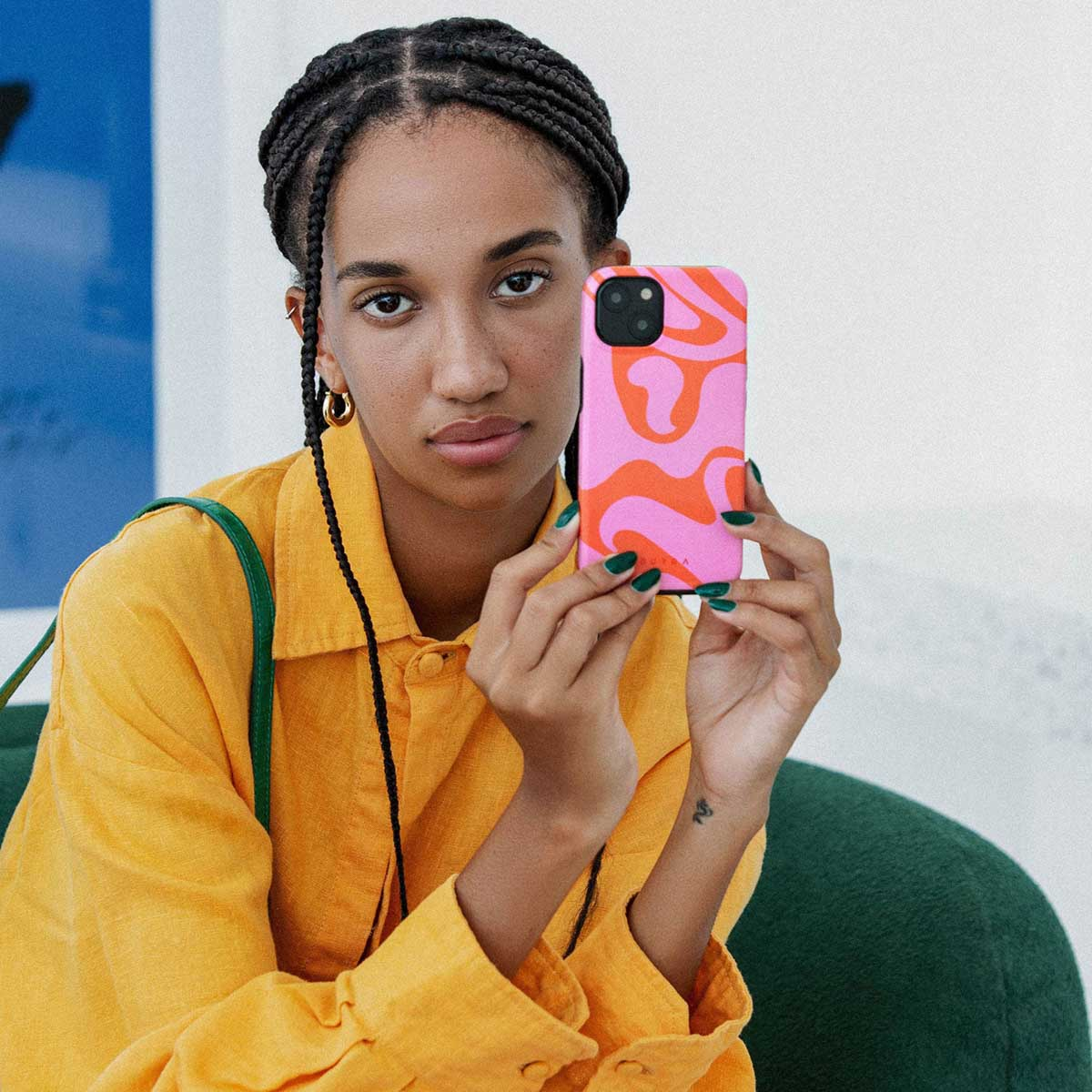 Een vrouw houdt een telefoon vast met een roze en oranje wervelcase. Ze draagt een gele blouse, gouden oorbellen en groene nagellak.