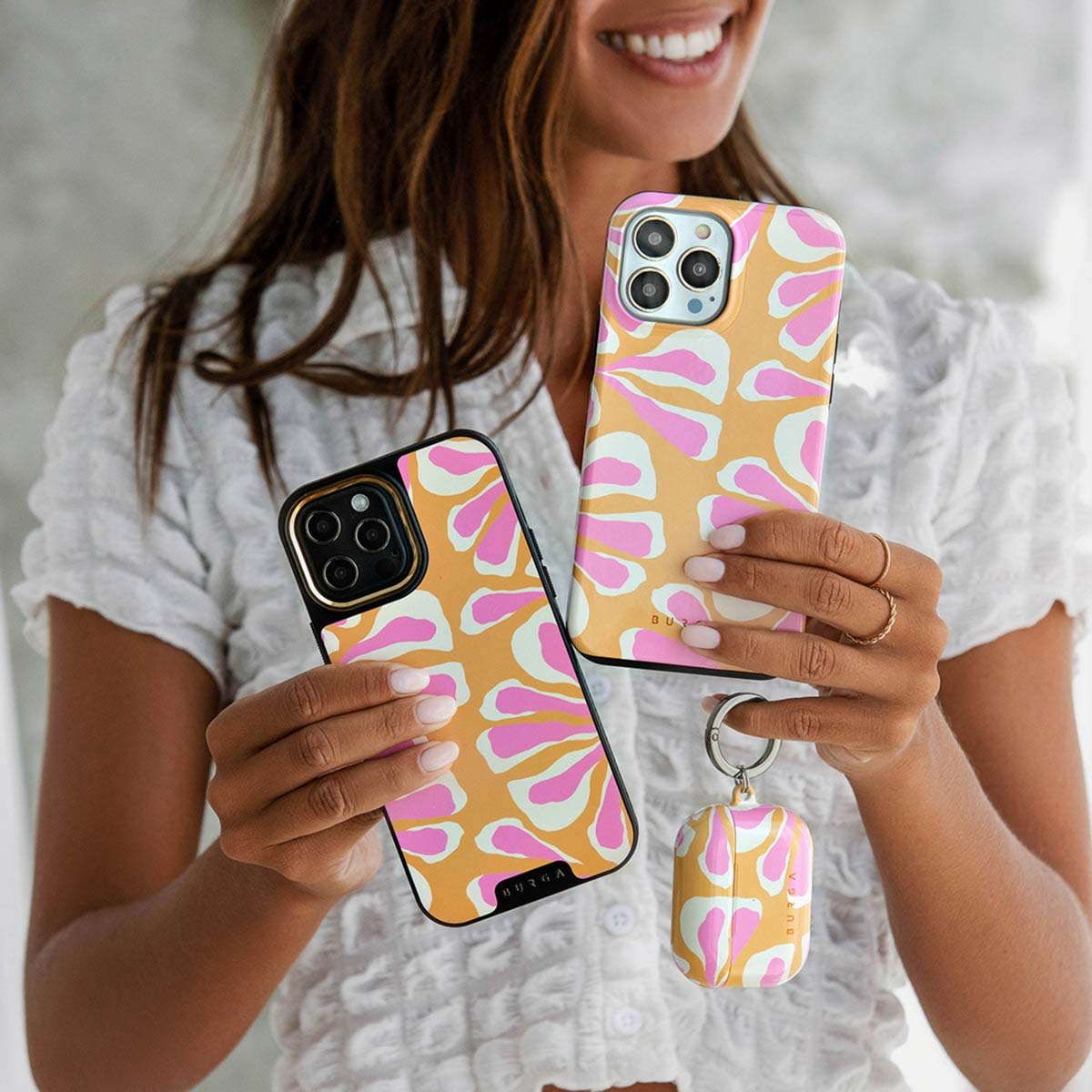 Een vrouw houdt twee telefoonhoesjes met patronen vast en een bijpassende AirPod-hoes. De hoesjes zijn oranje, roze en wit. Ze draagt een wit shirt.