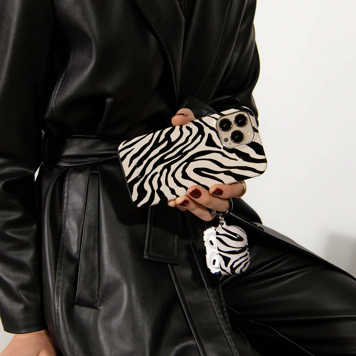 Een persoon houdt een telefoon en een AirPod-hoesje vast, beide met zebraprint. Ze draagt een zwarte leren jas. Rode nagels zijn zichtbaar.