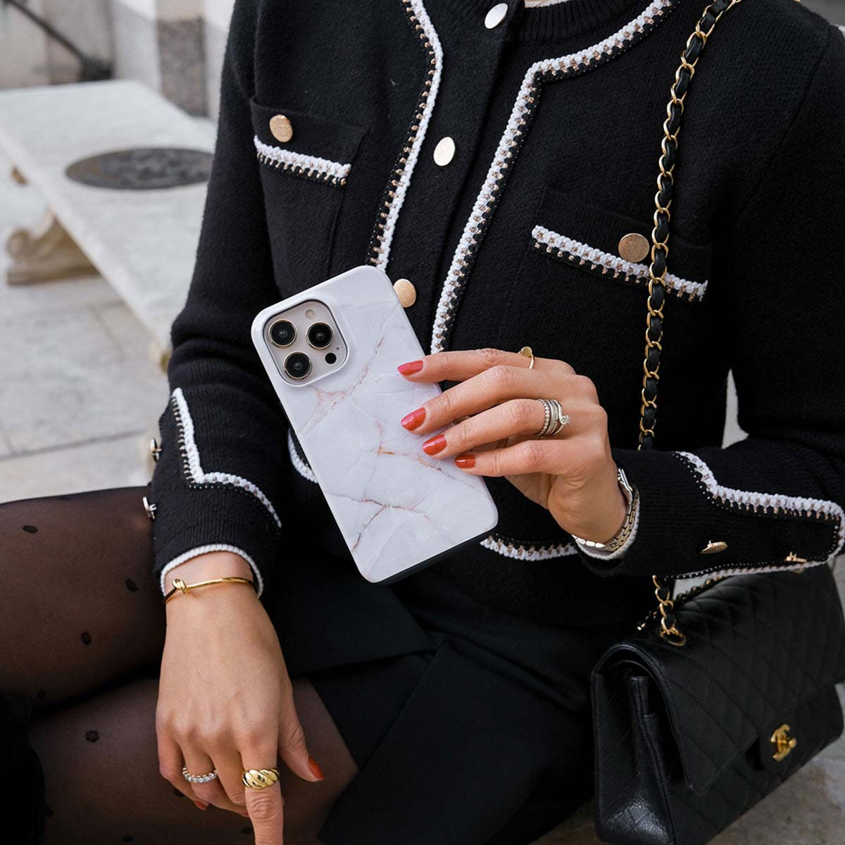 Vrouw met marmeren telefoonhoesje, draagt ​​zwarte jas, sieraden en een zwarte tas met gouden ketting.