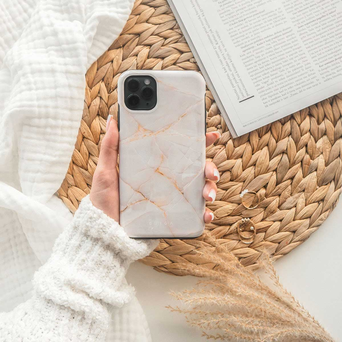 Een hand die een marmeren telefoonhoes vasthoudt, rust op een geweven mat, met een boek en ringen. Witte stof en gedroogde bloemen zijn te zien.