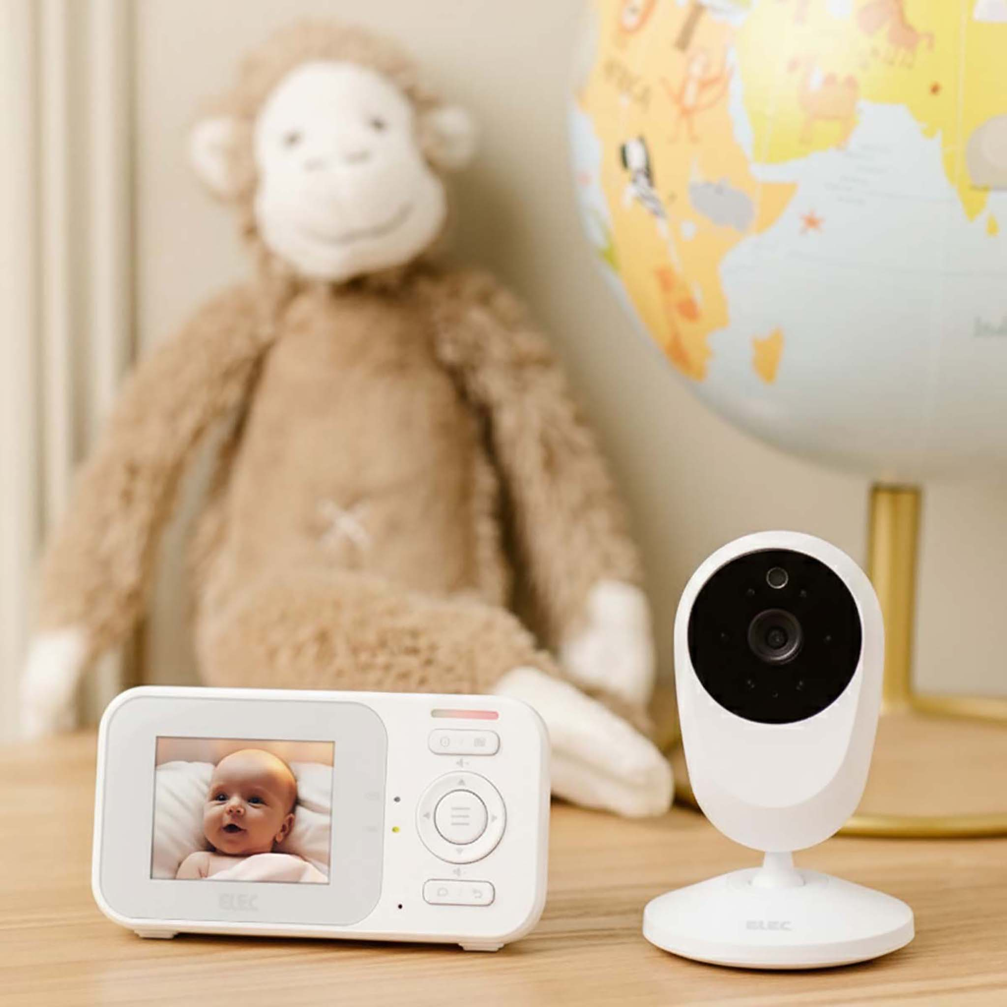 Ein Babyphone, ein Stoffaffe und ein Globus stehen auf einer Holzoberfläche. Der Monitor zeigt ein Baby.