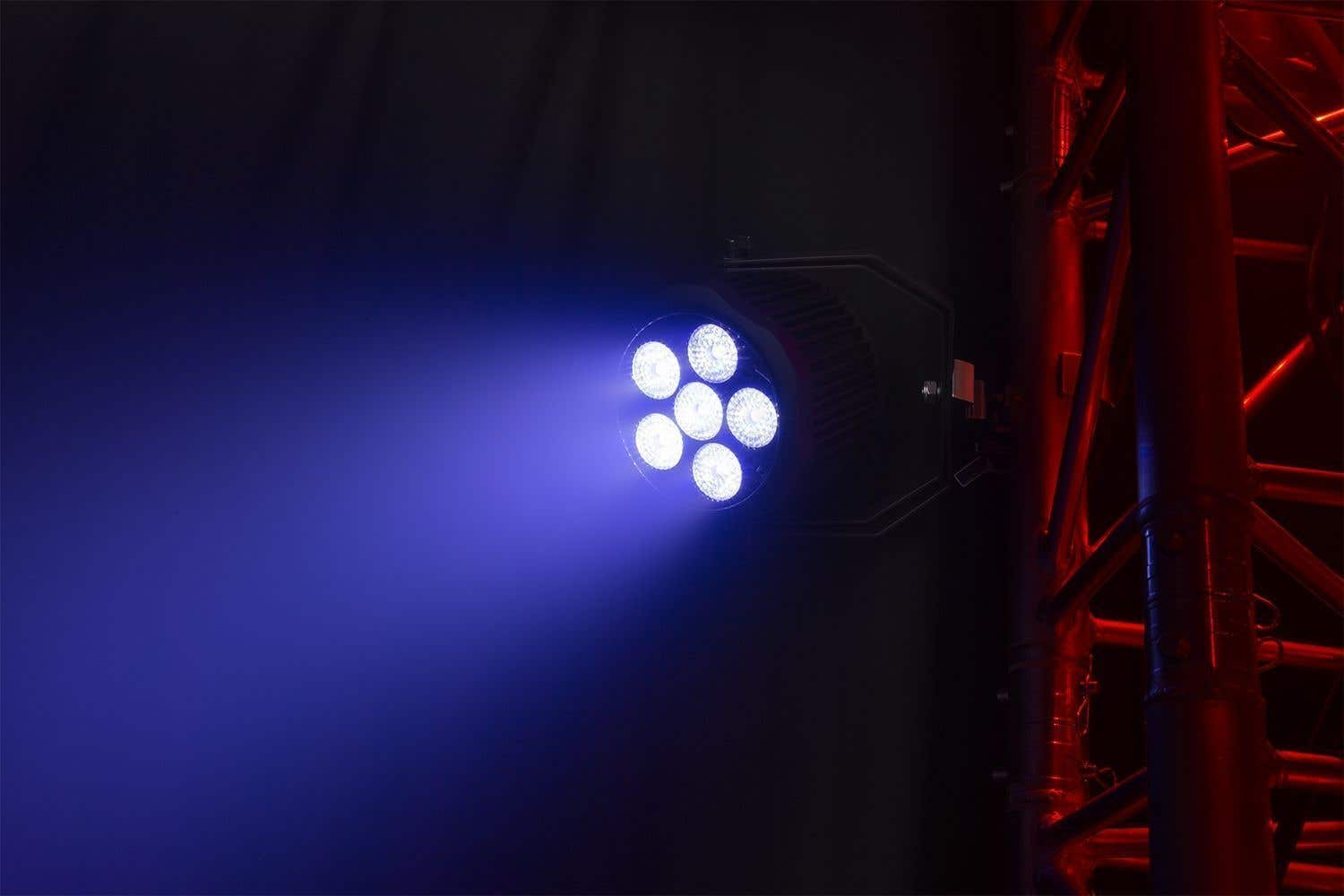 Foco escénico con siete bombillas brillantes que emiten un haz azul. Una estructura metálica roja se encuentra en el lado derecho del fondo oscuro.