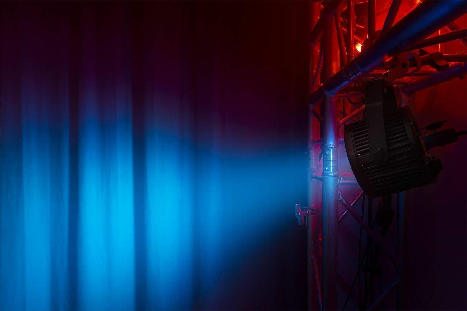 Una luz de escenario que ilumina una cortina con luz azul. Se ve una estructura metálica a la derecha, iluminada en rojo.