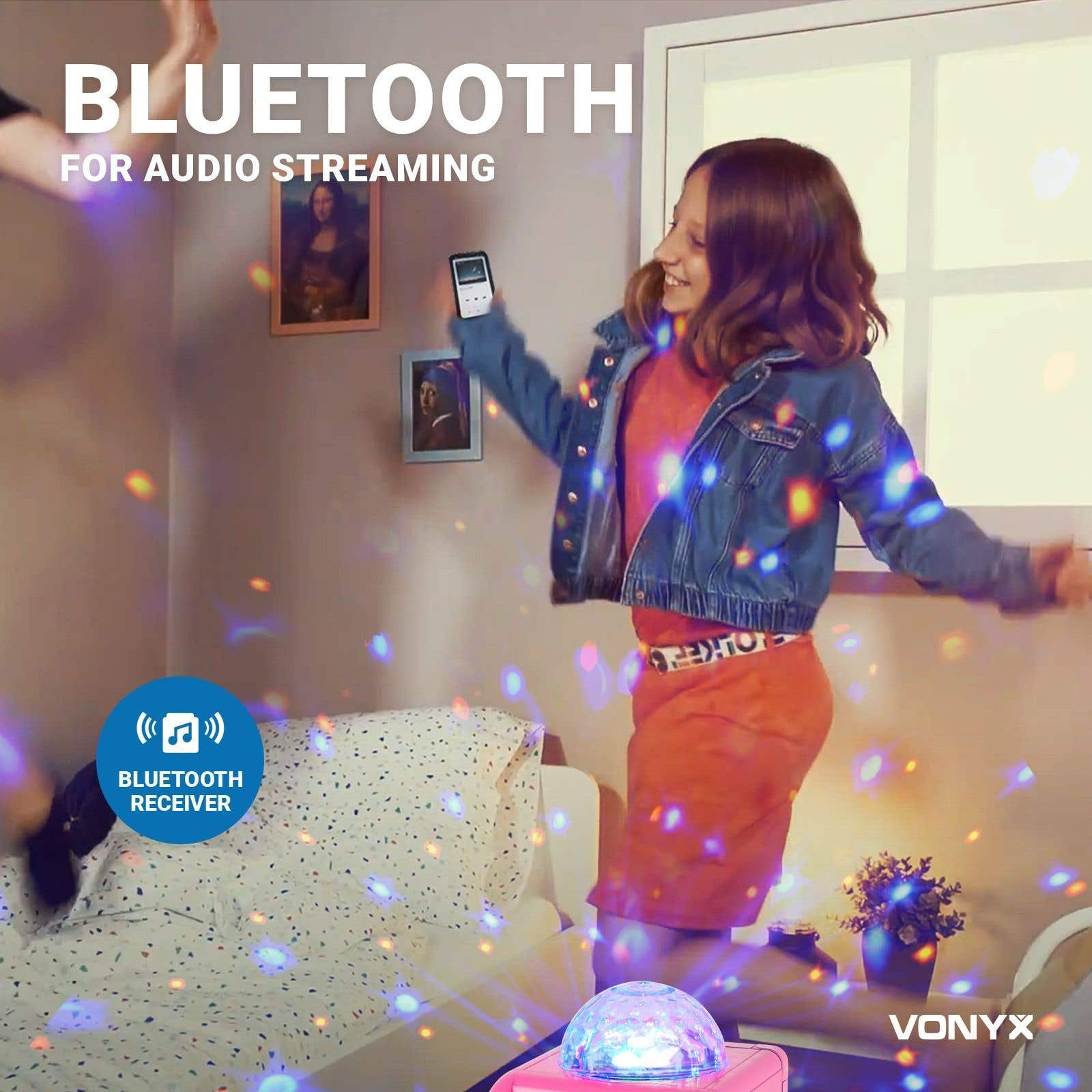Una niña baila con un teléfono. Una bola de discoteca proyecta luces. Receptor Bluetooth visible. Pinturas cuelgan en la pared.