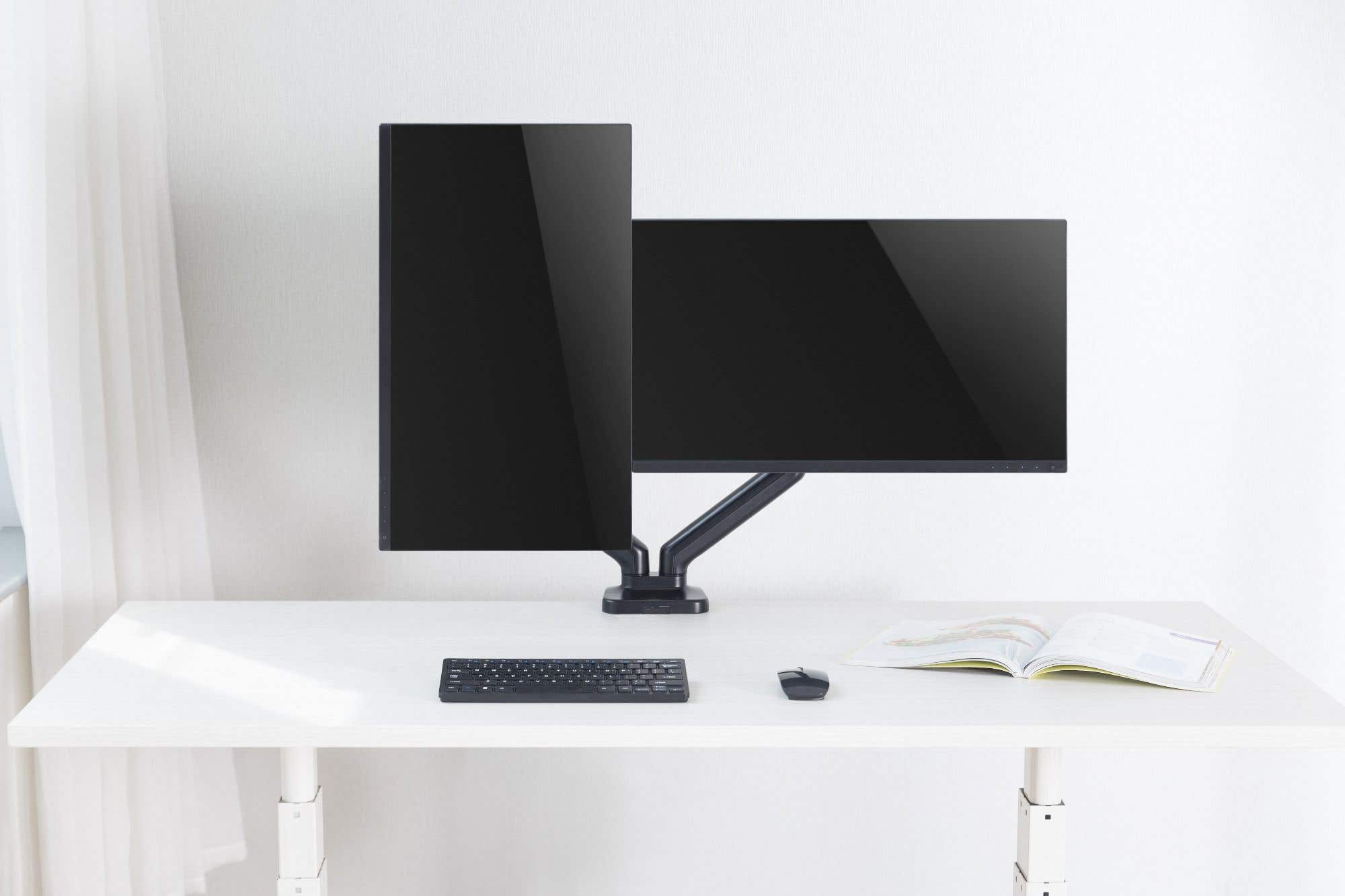 Two black computer monitors on a white desk. A keyboard, mouse, and open book are also on the desk. White wall in background.