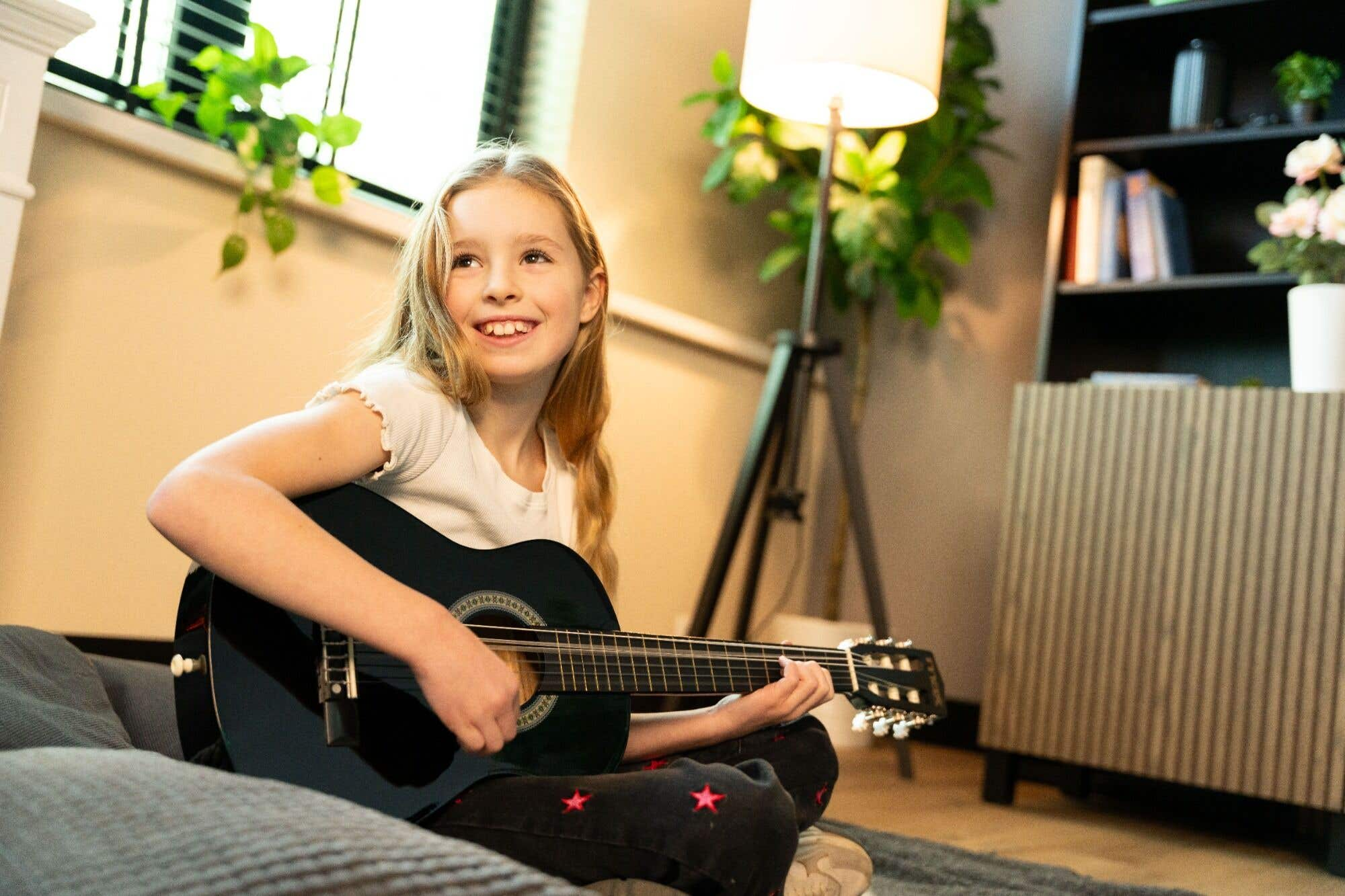 Een lachend meisje speelt gitaar. Ze zit op een bank. Lamp, raam en boekenkast op de achtergrond.