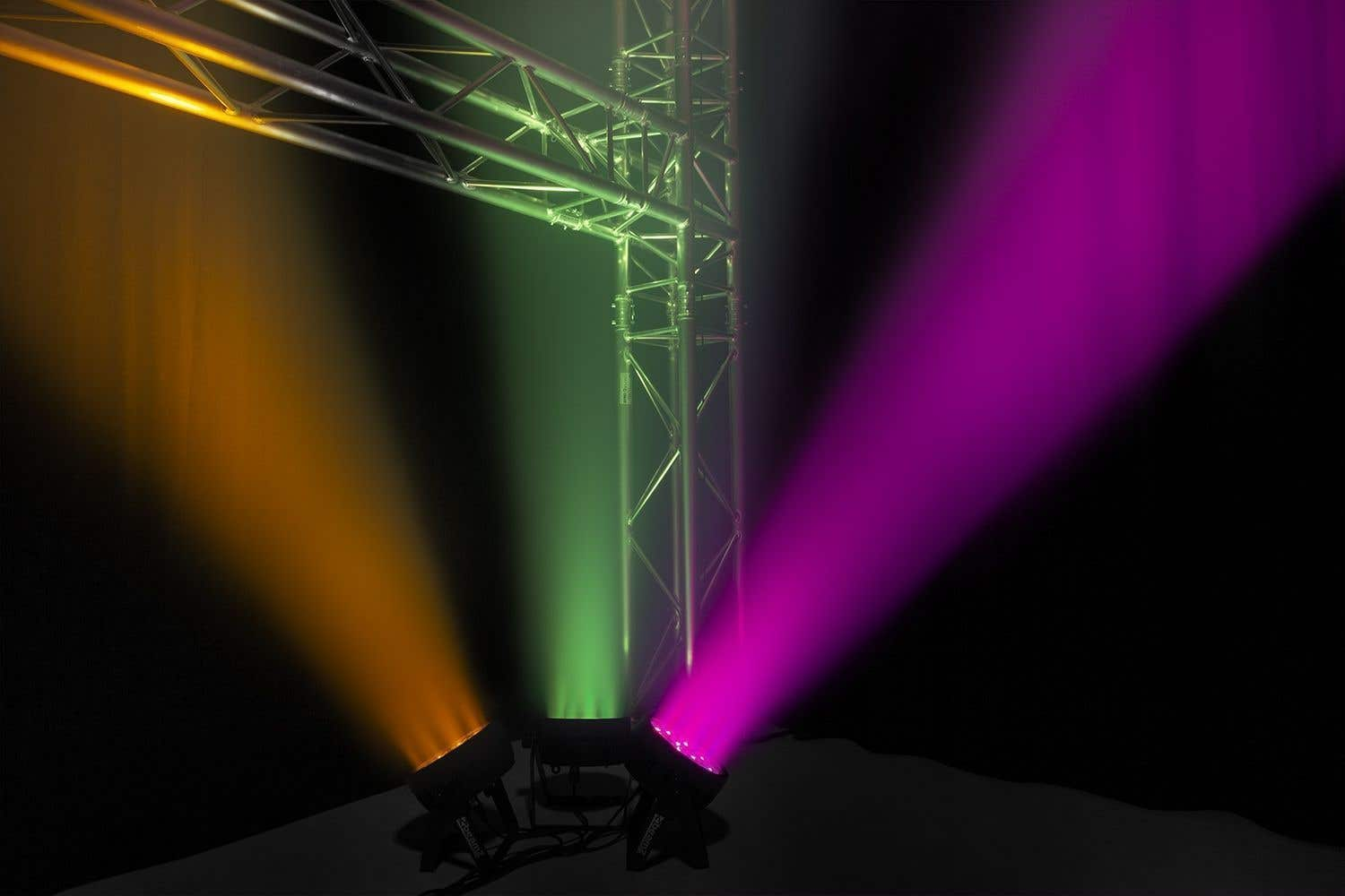 Luces de escenario proyectan haces naranjas, verdes y morados. Andamios metálicos están arriba y entre las luces. Fondo oscuro.