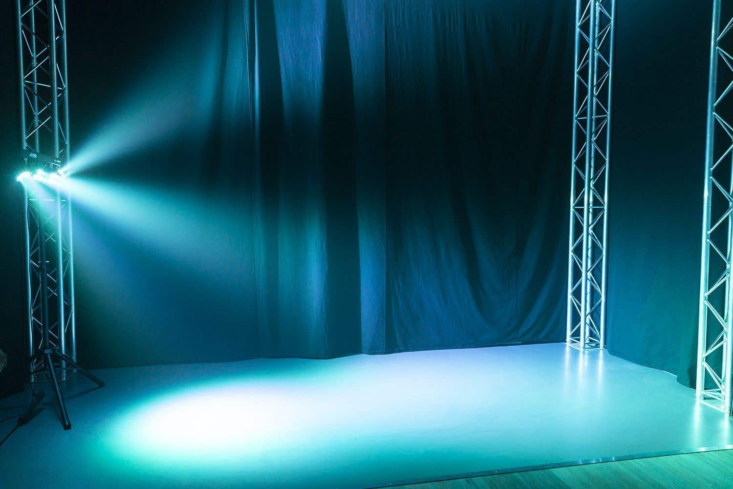 Un escenario iluminado con luces azules y verdes. Estructuras metálicas sostienen luces, proyectando haces sobre el suelo y el fondo oscuro.